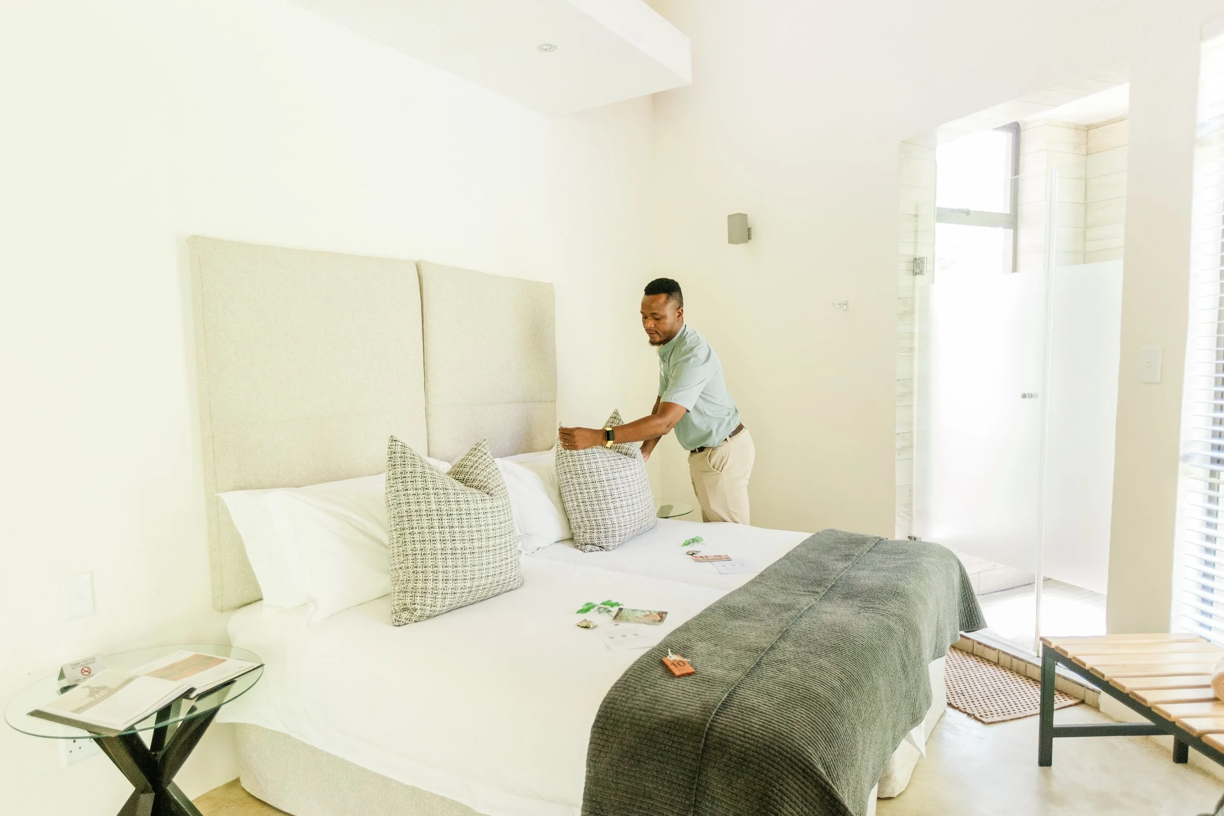 A man arranges decorative pillows on a bed in a bright, modern bedroom with large windows and minimalist decor.