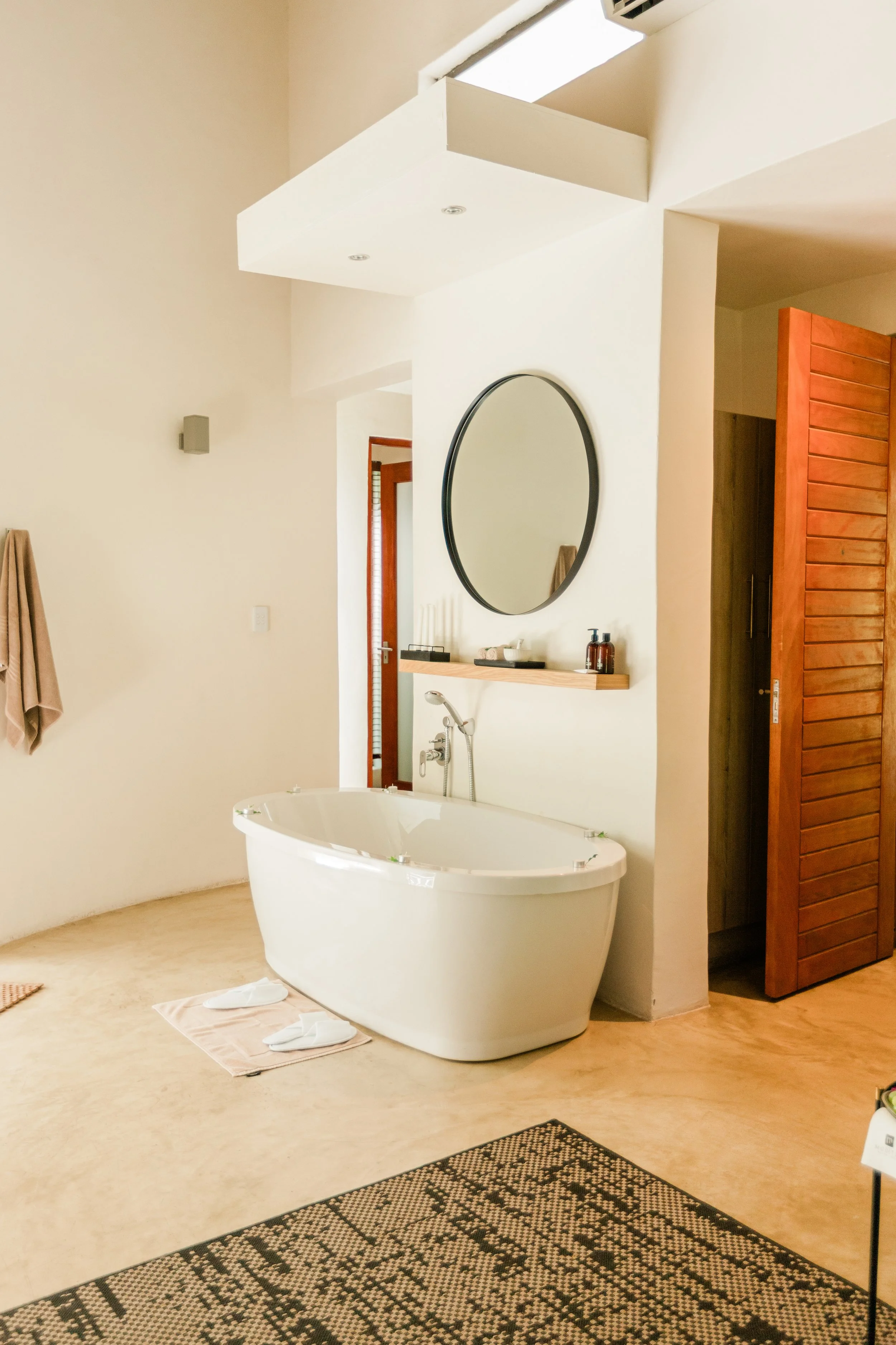 A bathroom with a large free-standing bathtub, a wall-mounted mirror, and a small wooden shelf with toiletries. The bathroom features a beige floor, a wooden door, and a window near the ceiling.