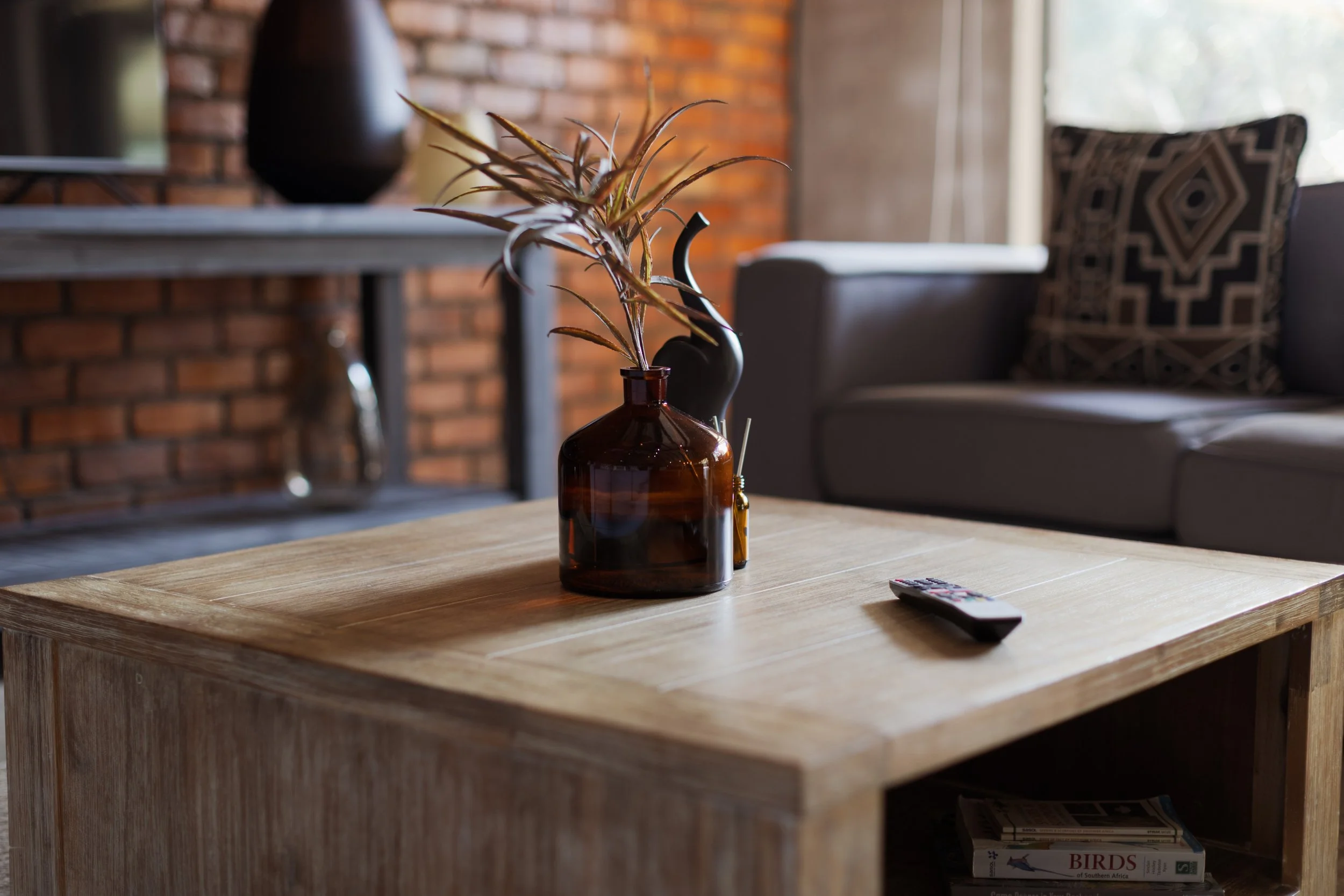 A brown glass vase with a narrow neck holding a dried plant arrangement on a wooden table in a living room. In the background, there is a dark vase, a window with sheer curtains, and a patterned pillow on a sofa.