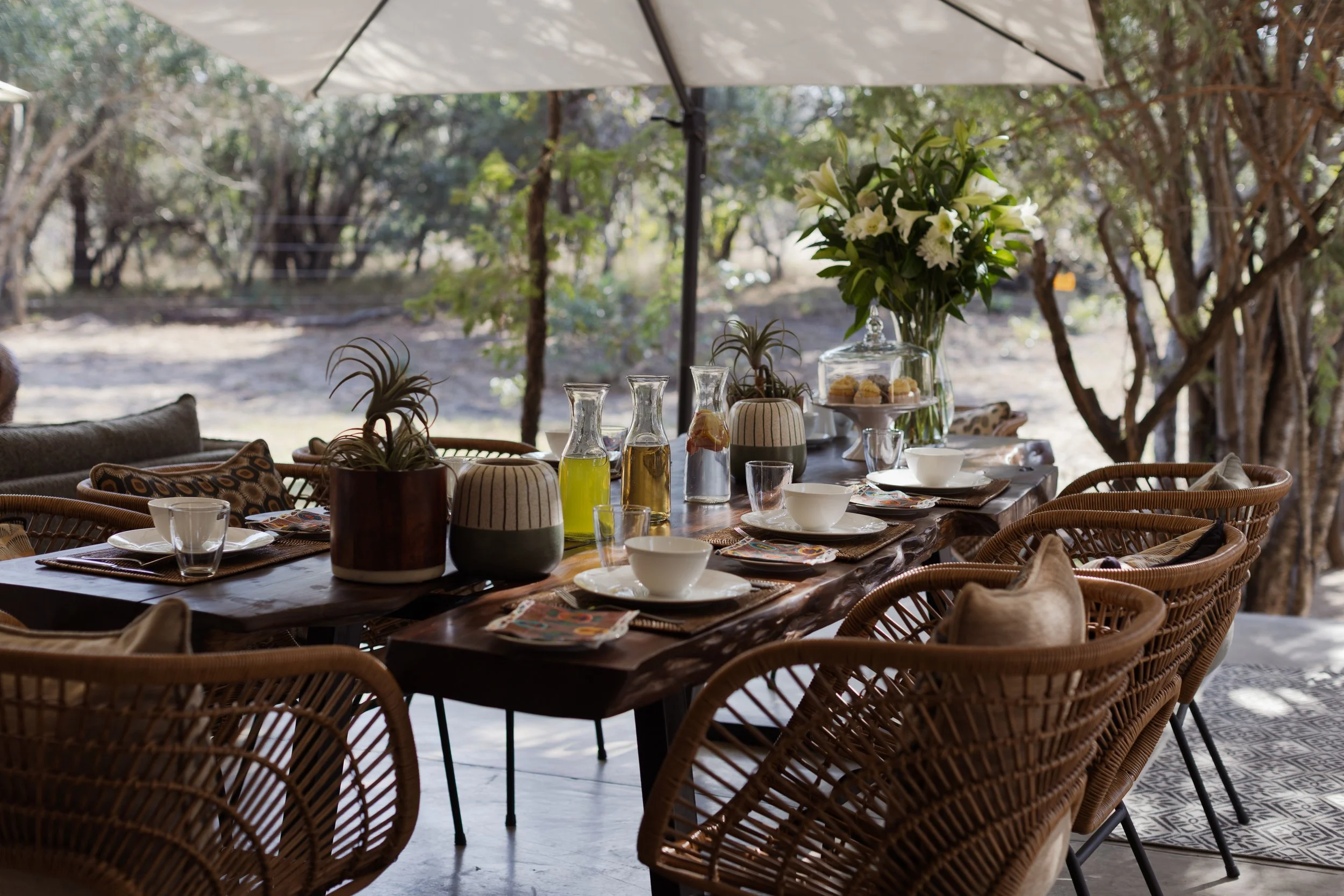 An outdoor dining setup with a large table, wicker chairs, and a white umbrella. The table is decorated with a vase of white flowers and various drinks, plates, and glassware.