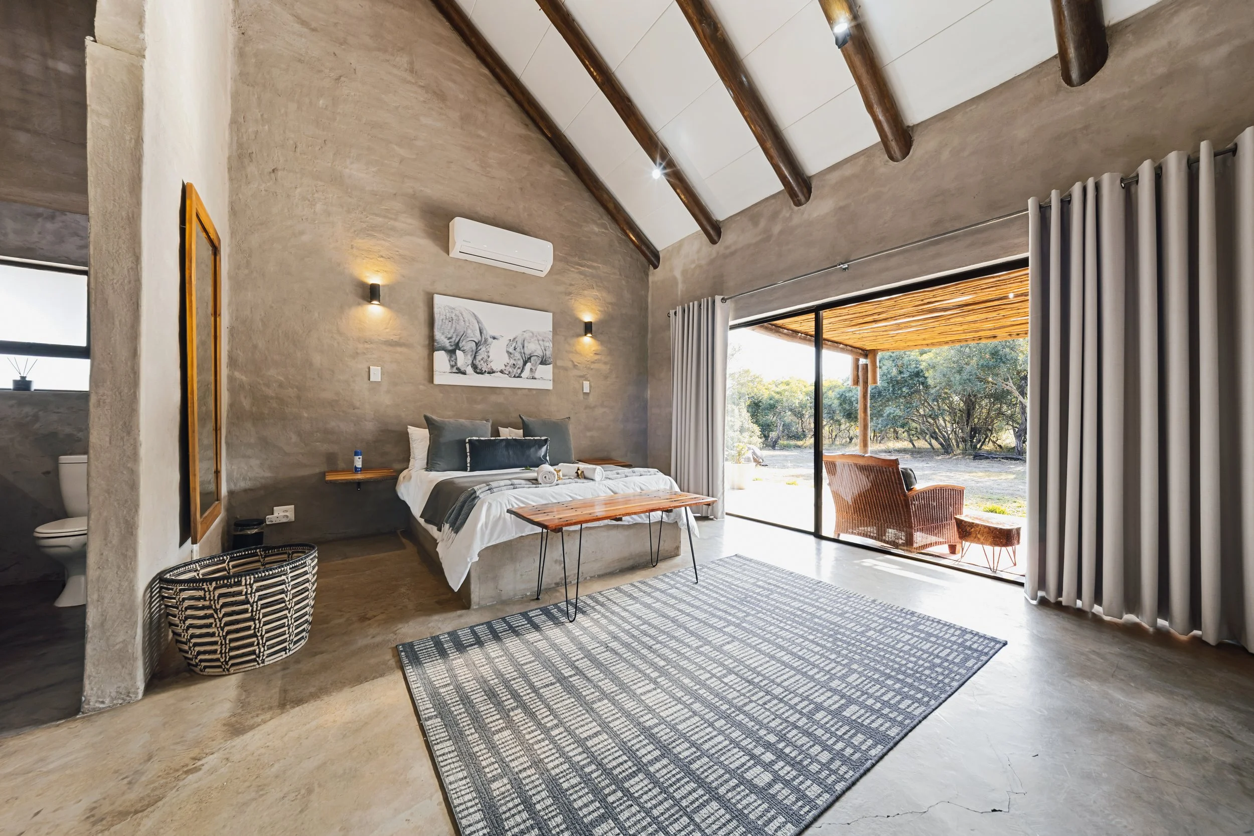 A modern bedroom with a sloped ceiling featuring wooden beams, concrete walls, a large sliding glass door with curtains leading to an outdoor patio, and minimalistic furniture including a bed, a bench, and chairs.