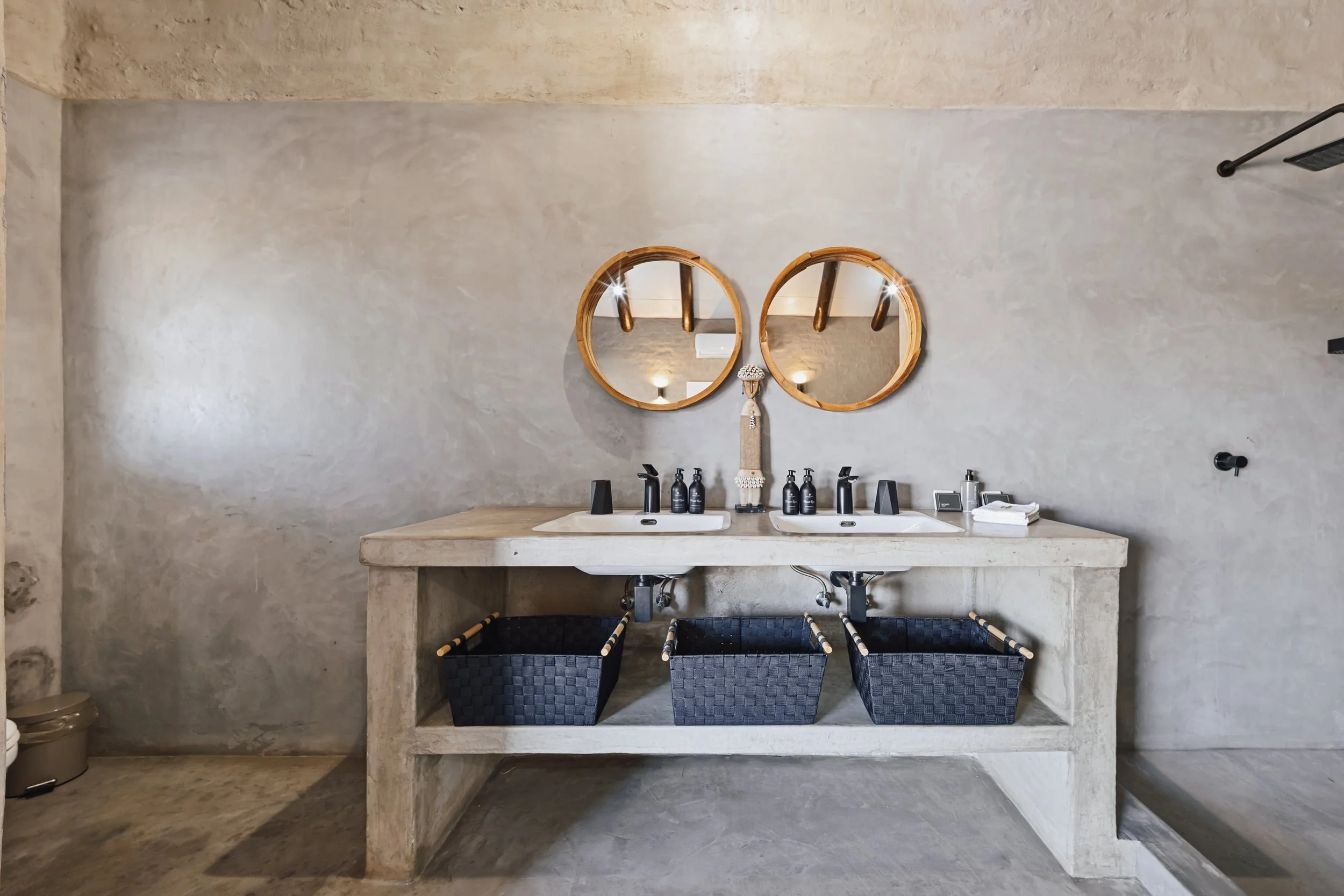 Bathroom vanity with double sinks, round wooden mirrors, black faucets, small decorative items, and black woven storage baskets underneath, in a rustic style with concrete surfaces.