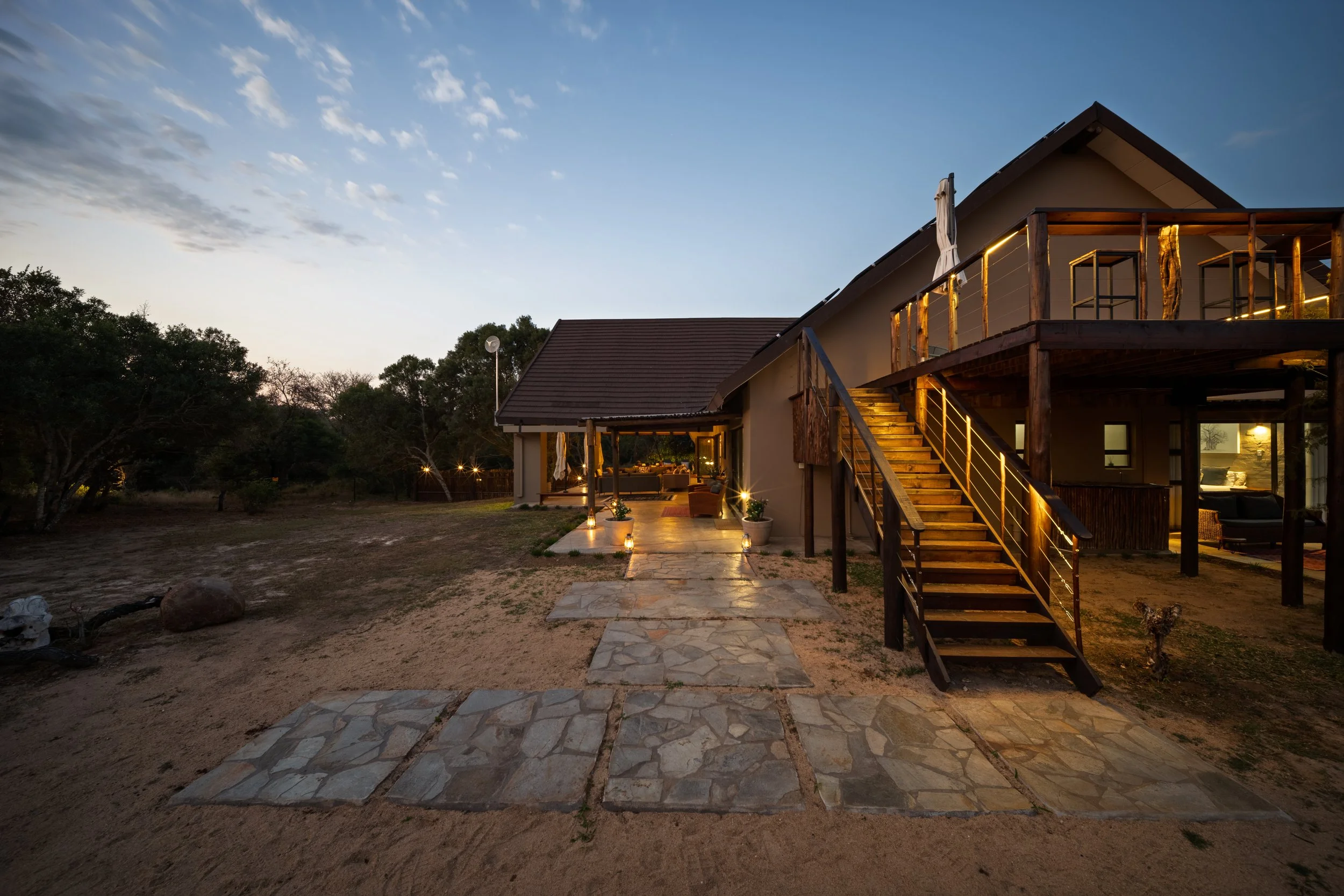 A two-story house with an outdoor staircase, glowing lights, and a patio at dusk. The house has a sloped roof and wooden accents, surrounded by trees and open land.