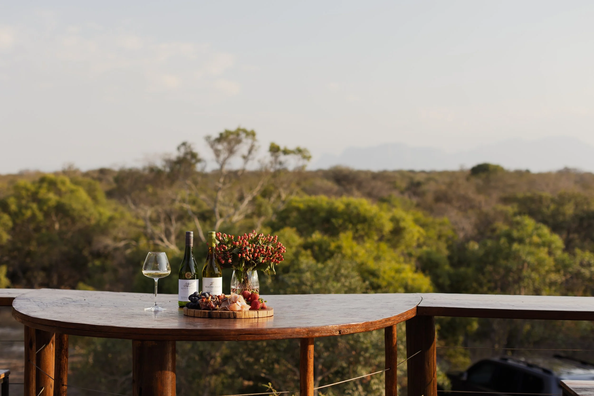 Outdoor table with wine bottles, wine glass, cheese, grapes, and flowers, set against a background of trees and hills.