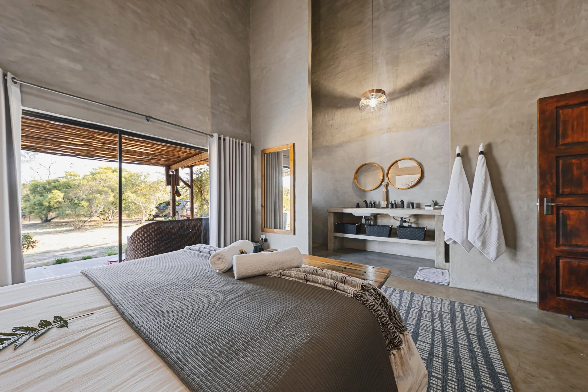A bedroom with a large bed, rolled towels, and decorative items. There is an open sliding door leading outside to trees and outdoors. The room has at least two sinks with mirrors and hanging towels, a wooden door, and a ceiling light, all with a mode