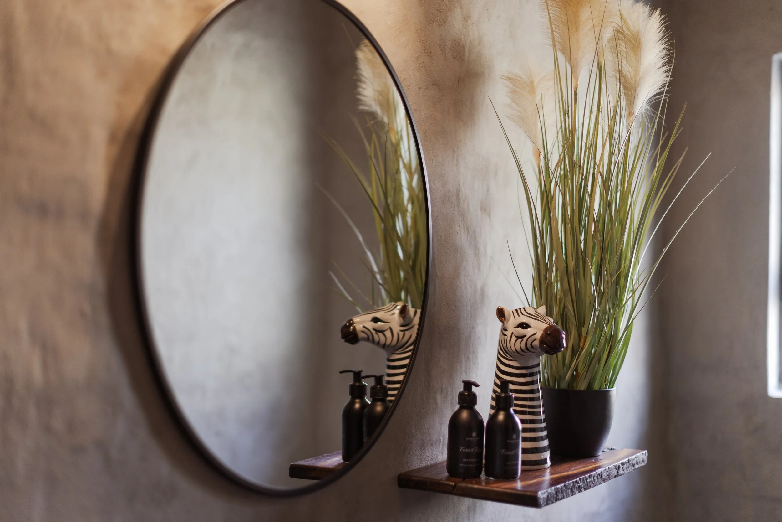 A round mirror hanging on a beige wall reflects a decorative zebra-striped vase filled with tall, wispy beige grass. On a wooden shelf below the mirror, there are two black bottles labeled 'Forest' and 'Champs,' positioned next to the vase.
