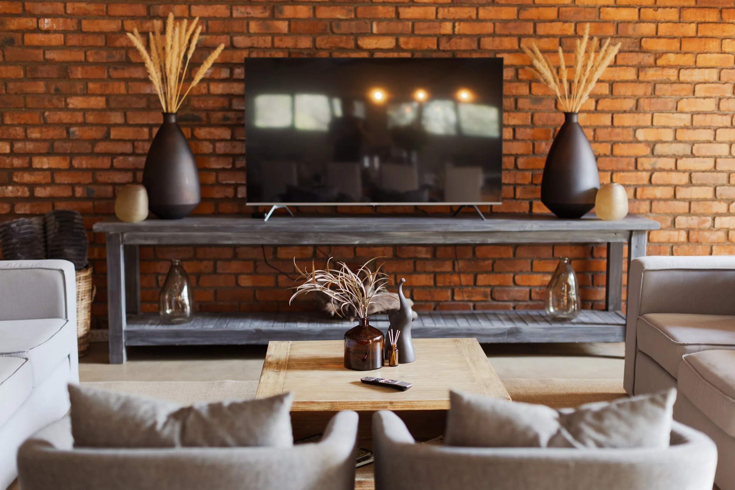 Living room with brick wall, TV on a wooden console table, and beige armchairs with a wooden coffee table in the center. Decor includes vases and dried plants.