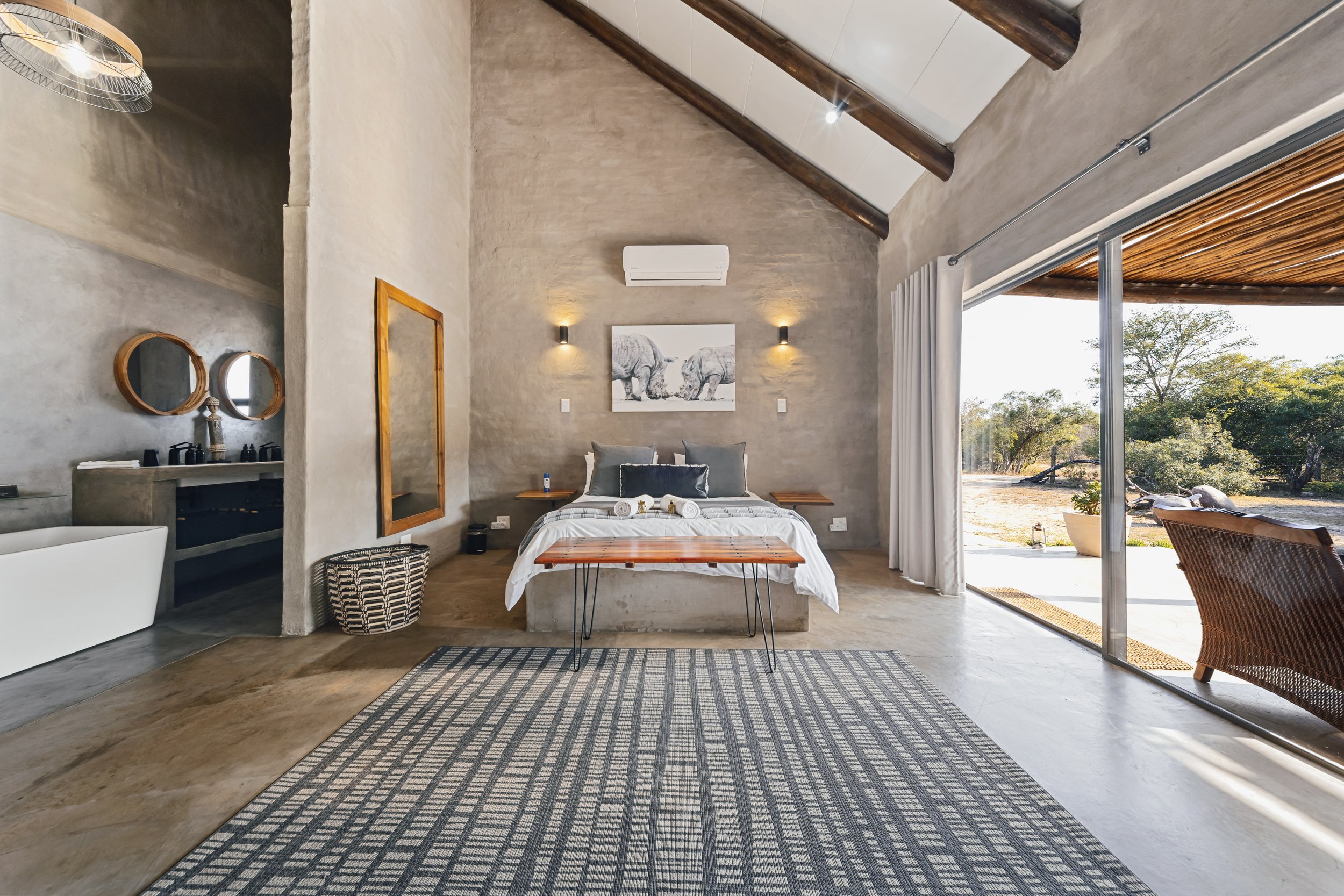 A modern bedroom with a high, sloped ceiling and large sliding glass door opening to an outdoor area with trees. The room features a bed with gray pillows, wall-mounted lights, a framed animal art print, and a patterned rug. Wooden beams and a small 