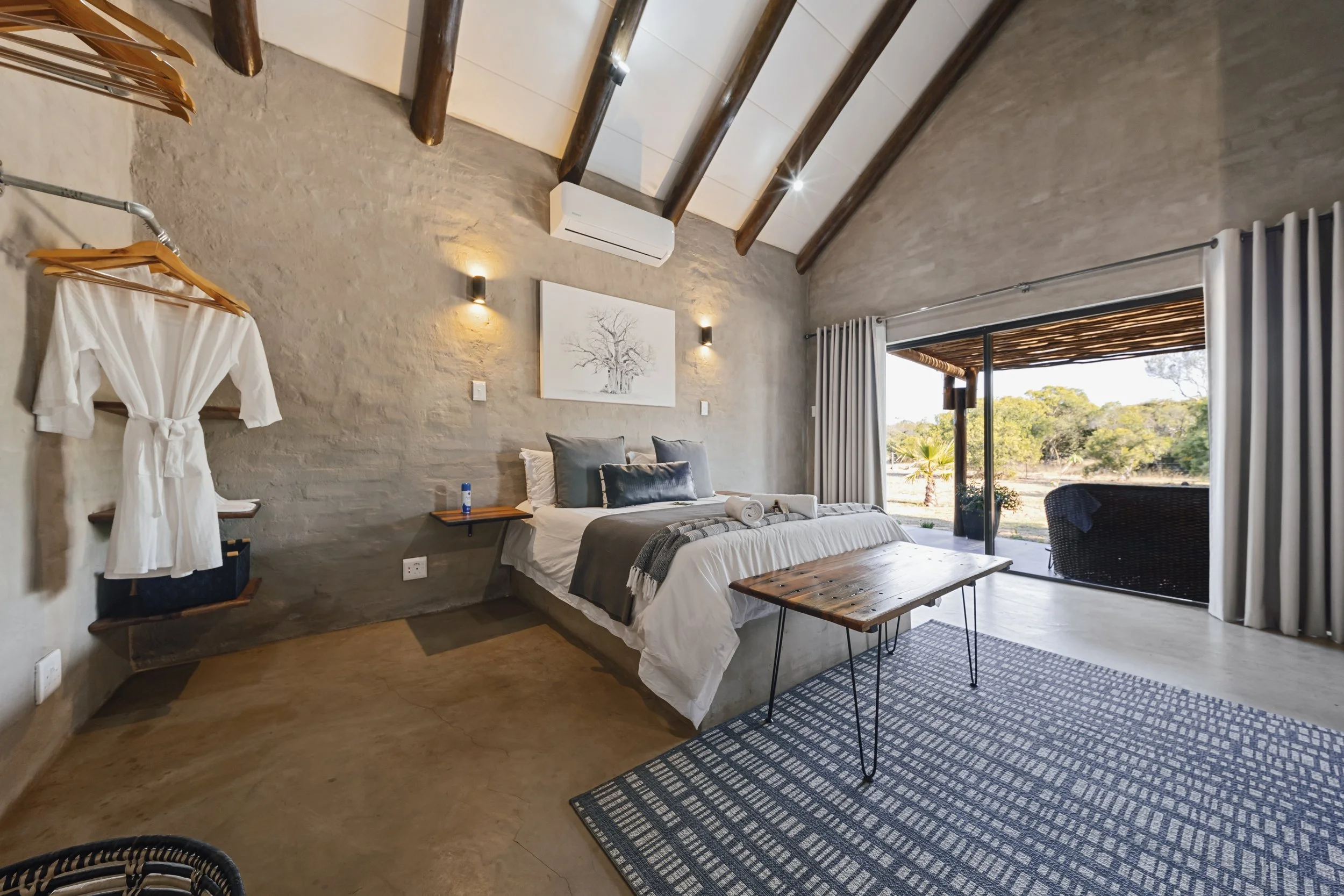 Modern bedroom with exposed wood ceiling beams, a stone accent wall, a large bed with gray and white bedding, a small wooden table, a black patterned area rug, and floor-to-ceiling sliding glass doors with white curtains opening to an outdoor view wi