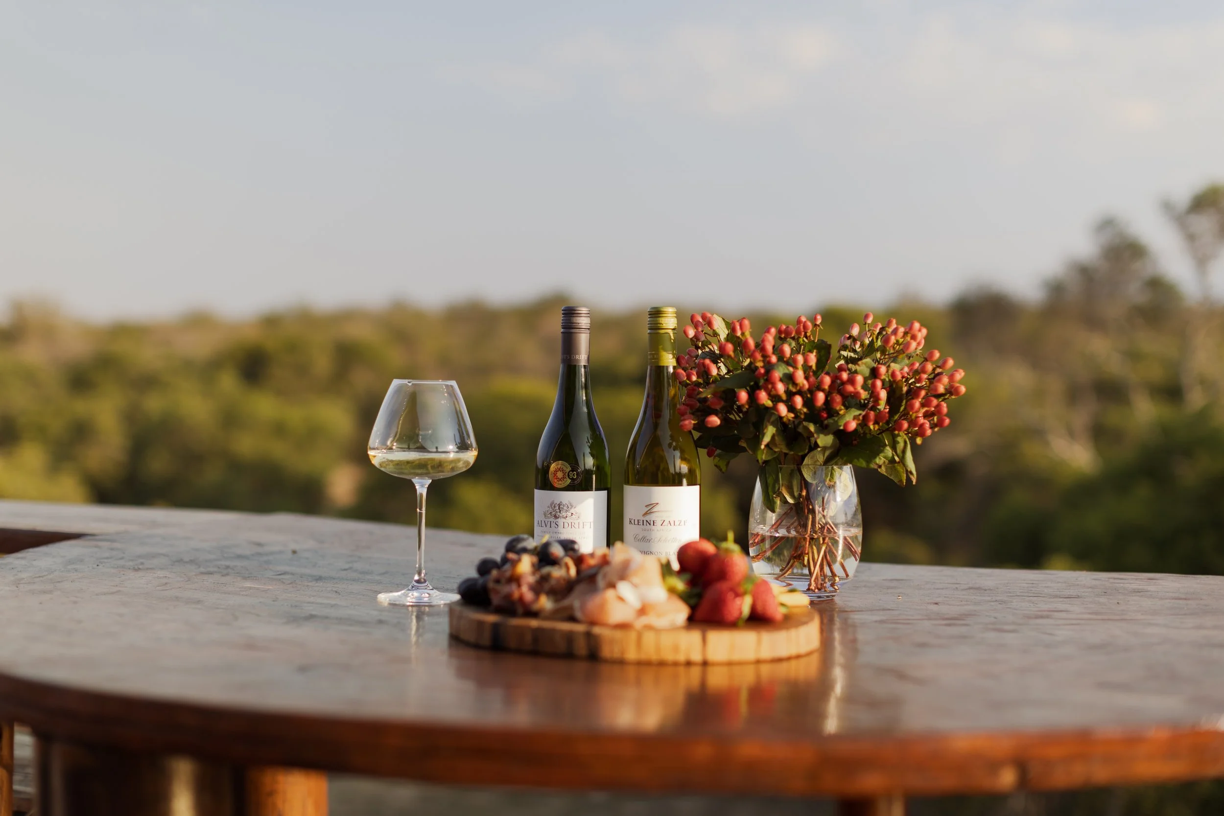 A table with wine bottles, a wine glass, a floral arrangement with red berries, and assorted fruits on a wooden platter set outdoors with a blurred natural landscape in the background.