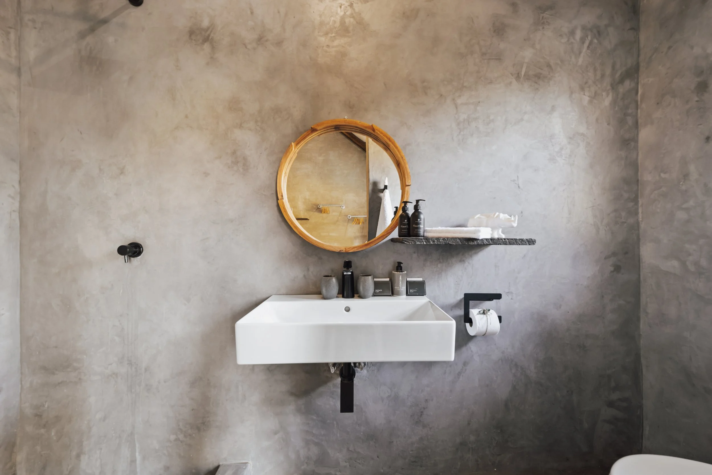 Bathroom with rectangular sink, black faucet and round wooden mirror on a concrete wall. 
