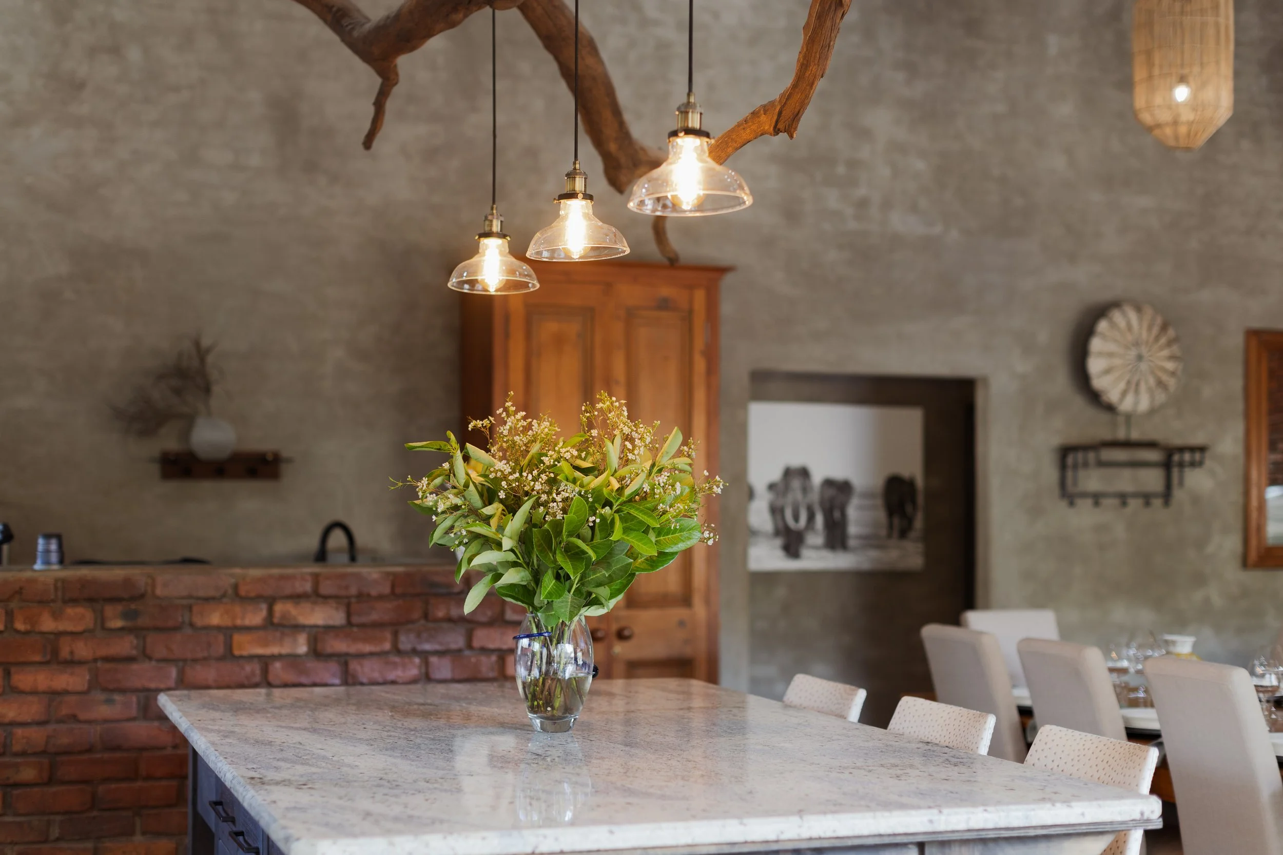 A glass vase with green leaves and small white flowers on a light-colored marble kitchen island in a rustic dining room with exposed brick, wooden cabinet, and decorative wall art.