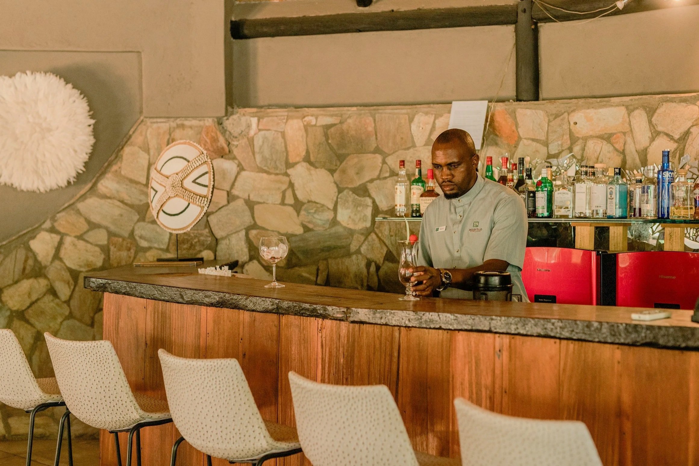 A bartender or server with a dark complexion, shaved head, and wearing a light-colored shirt, stands behind a wooden bar with a glass of drink in hand, in front of a stone wall with various bottles of alcohol.