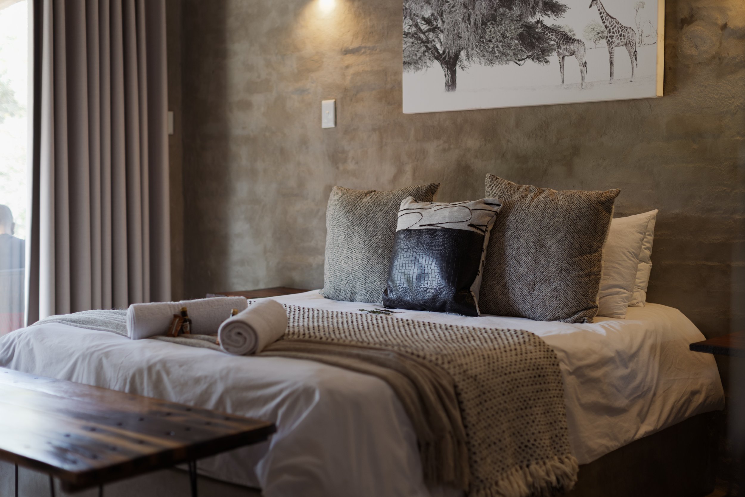 A neatly made bed with pillows and a rolled towel on top, in a bedroom with concrete walls, a large window with curtains, and a giraffe artwork on the wall.
