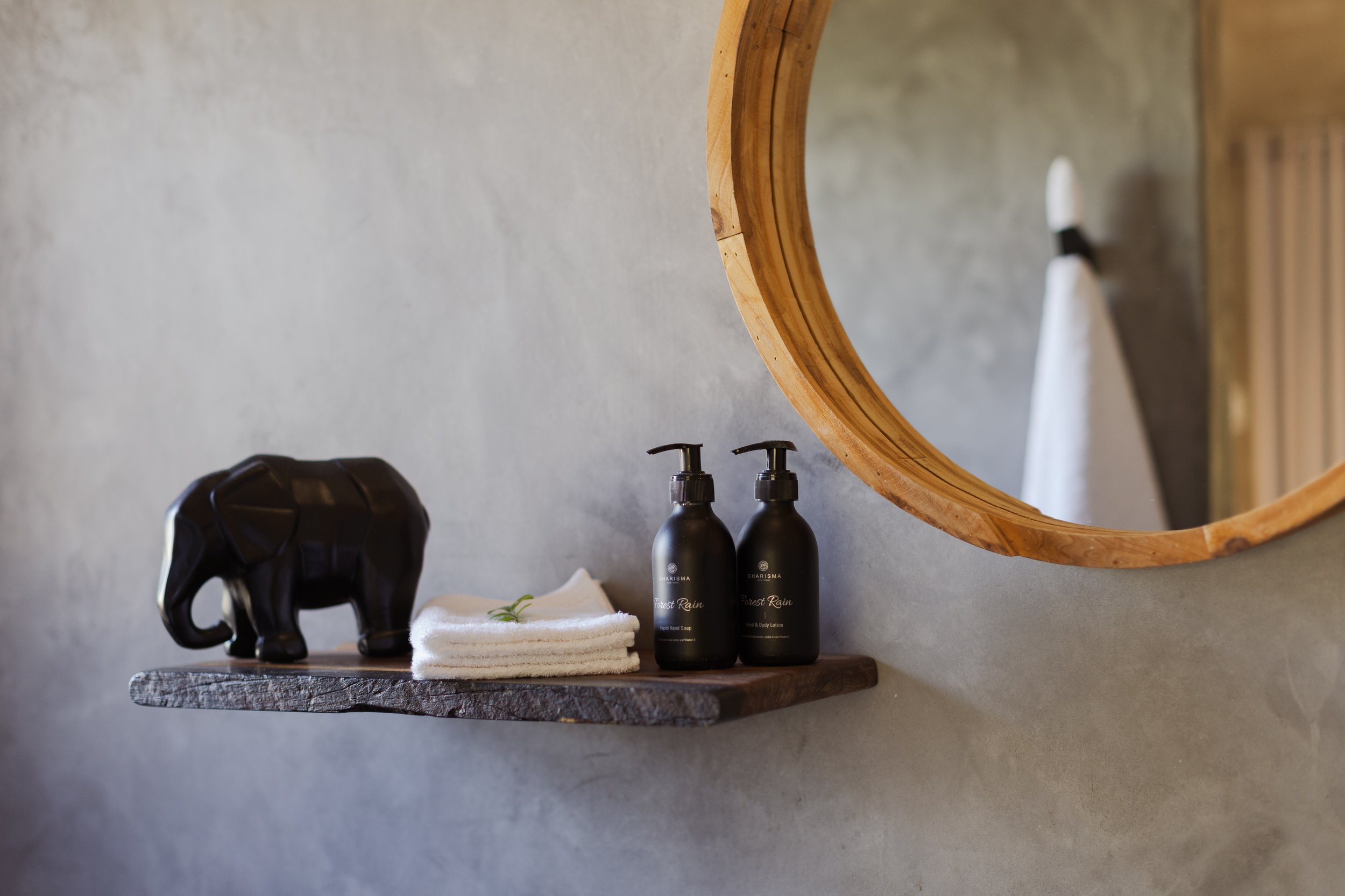 A wooden shelf holding a black decorative elephant sculpture, two black bottles labeled 'Forest Rain' hand soap and lotion, and a rolled white towel with a sprig of green herbs. A circular mirror with a wooden frame and a hand towel are visible in th