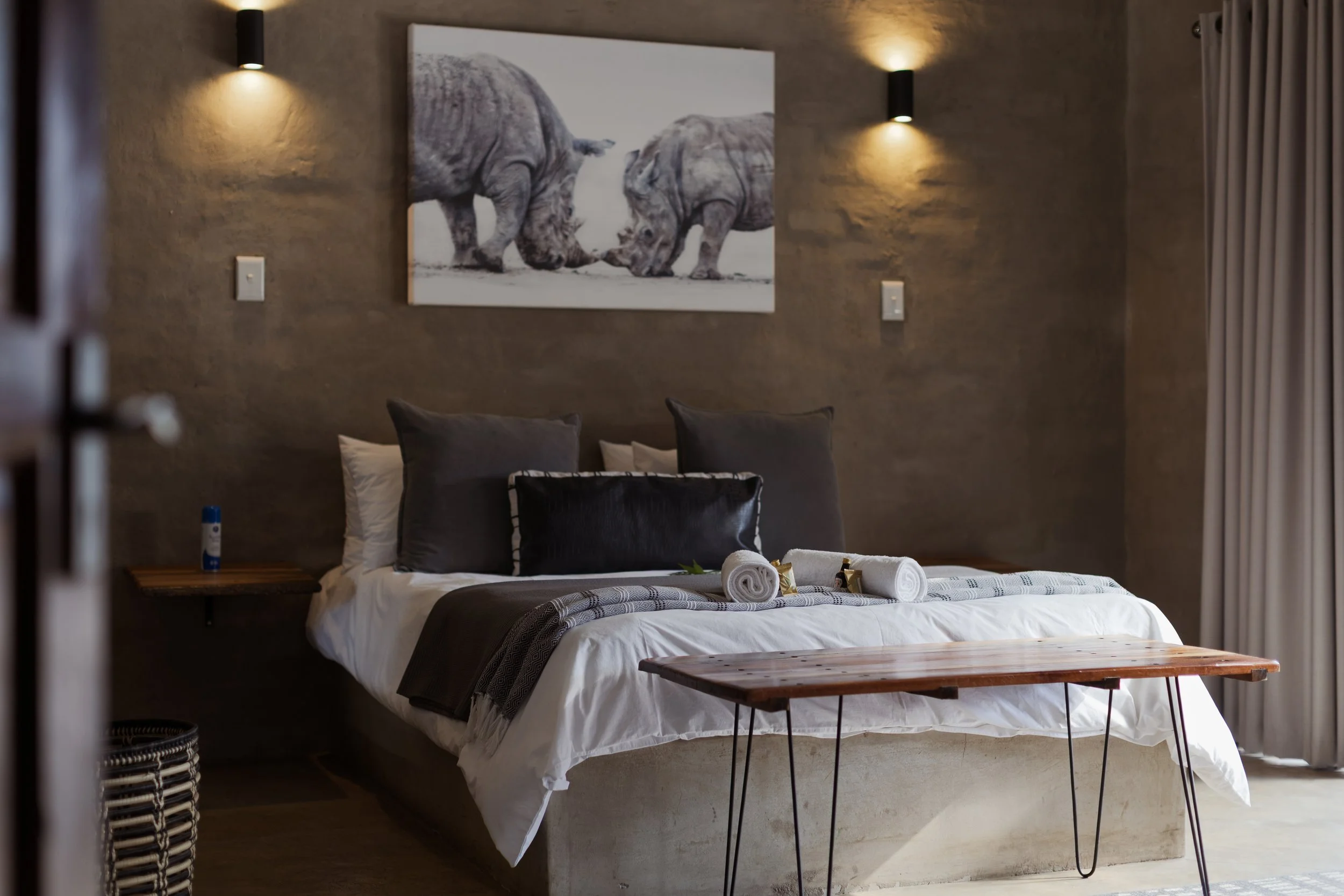 A neatly made bed with white sheets, dark pillows, and rolled towels on top. Behind the bed, there is a large artwork of two elephants facing each other. The room has a textured beige wall with two wall-mounted lights and curtains on the right side.
