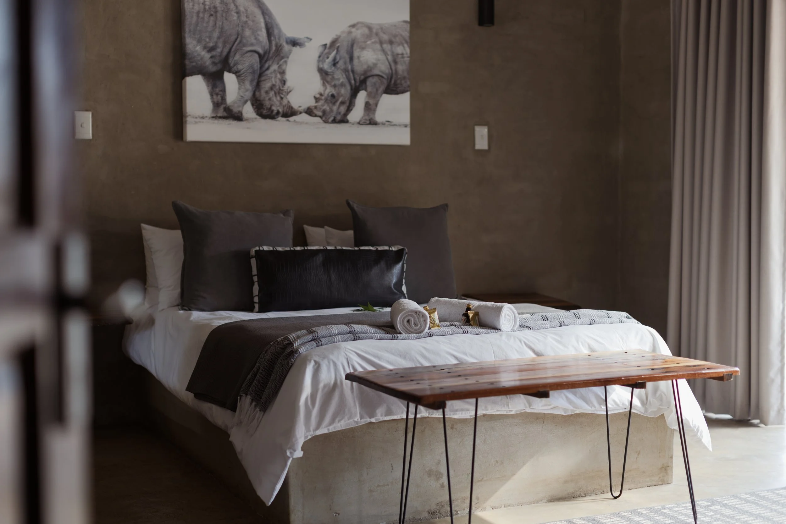 A neatly made bed with white sheets, gray pillows, and rolled towels on top, with a wooden bench at the foot of the bed. Above the bed hangs a picture of two rhinoceroses touching noses. The room has beige walls, a light-colored carpet, and curtains 