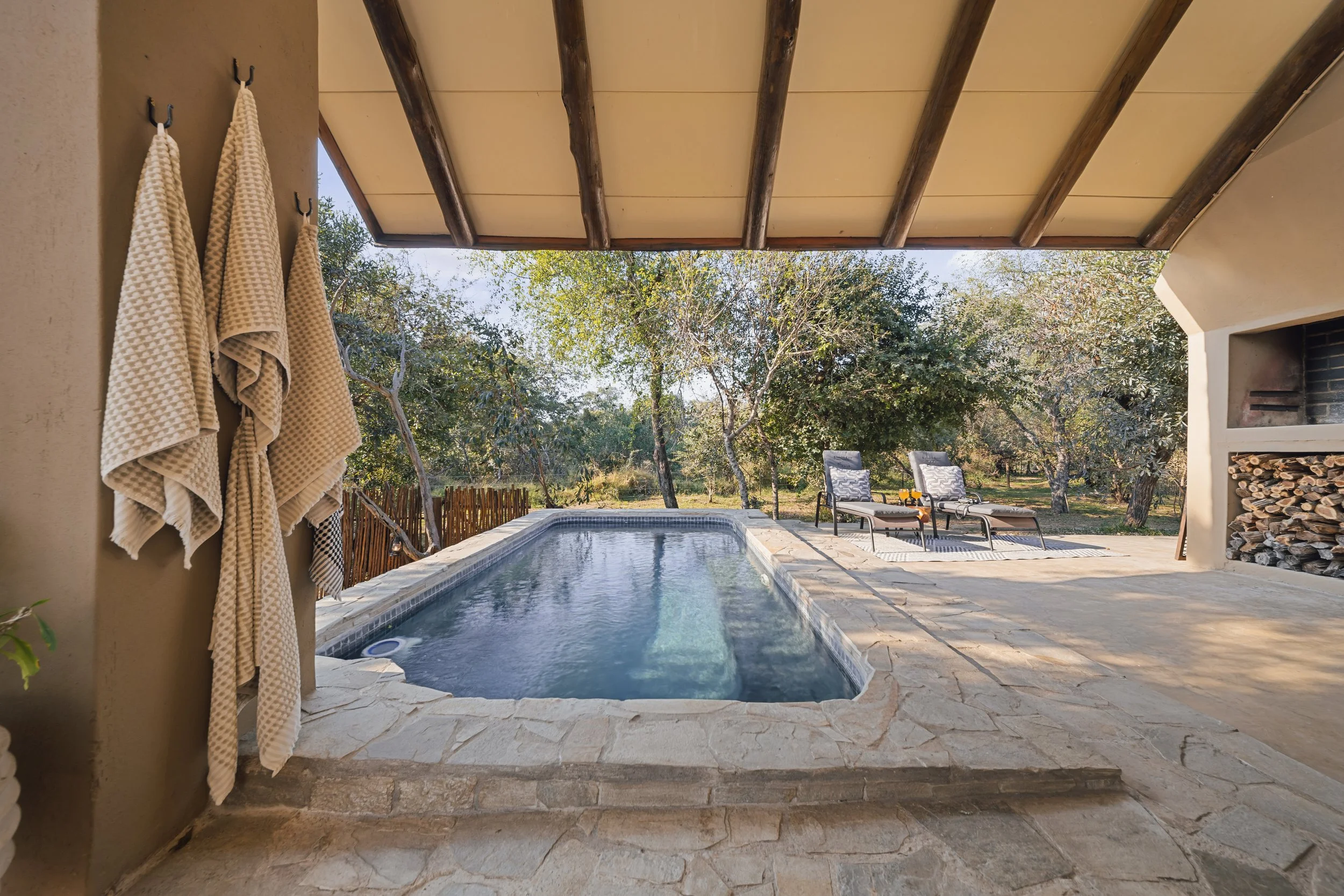 A backyard scene with a small pool, lounge chairs, and a stacked wood firepit, surrounded by trees and outdoors.