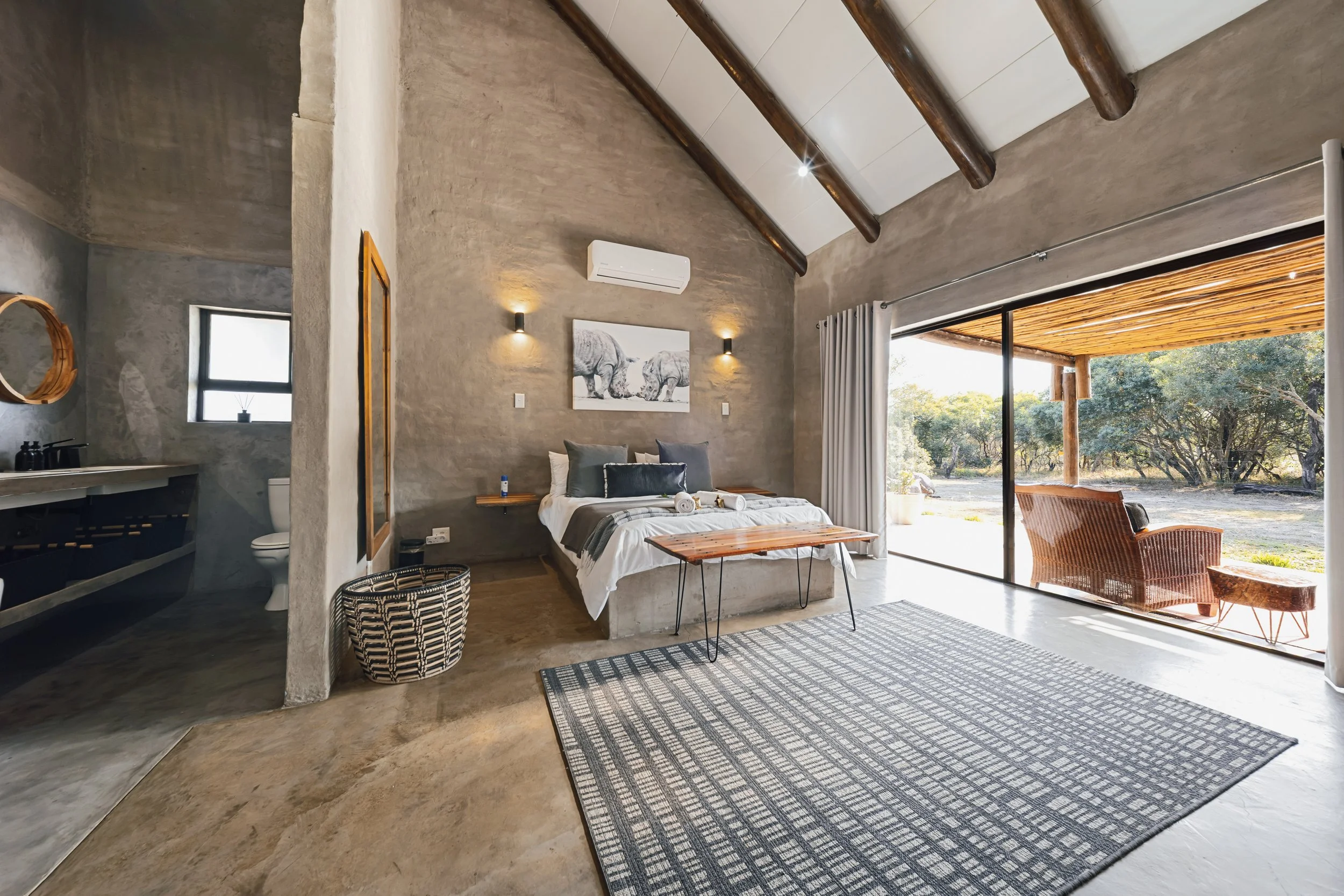 A modern bedroom with high vaulted ceiling supported by dark wooden beams, a large sliding glass door leading to an outdoor area, a bed with gray and white bedding, a black and white patterned rug, and minimal decor including a picture of two elephan