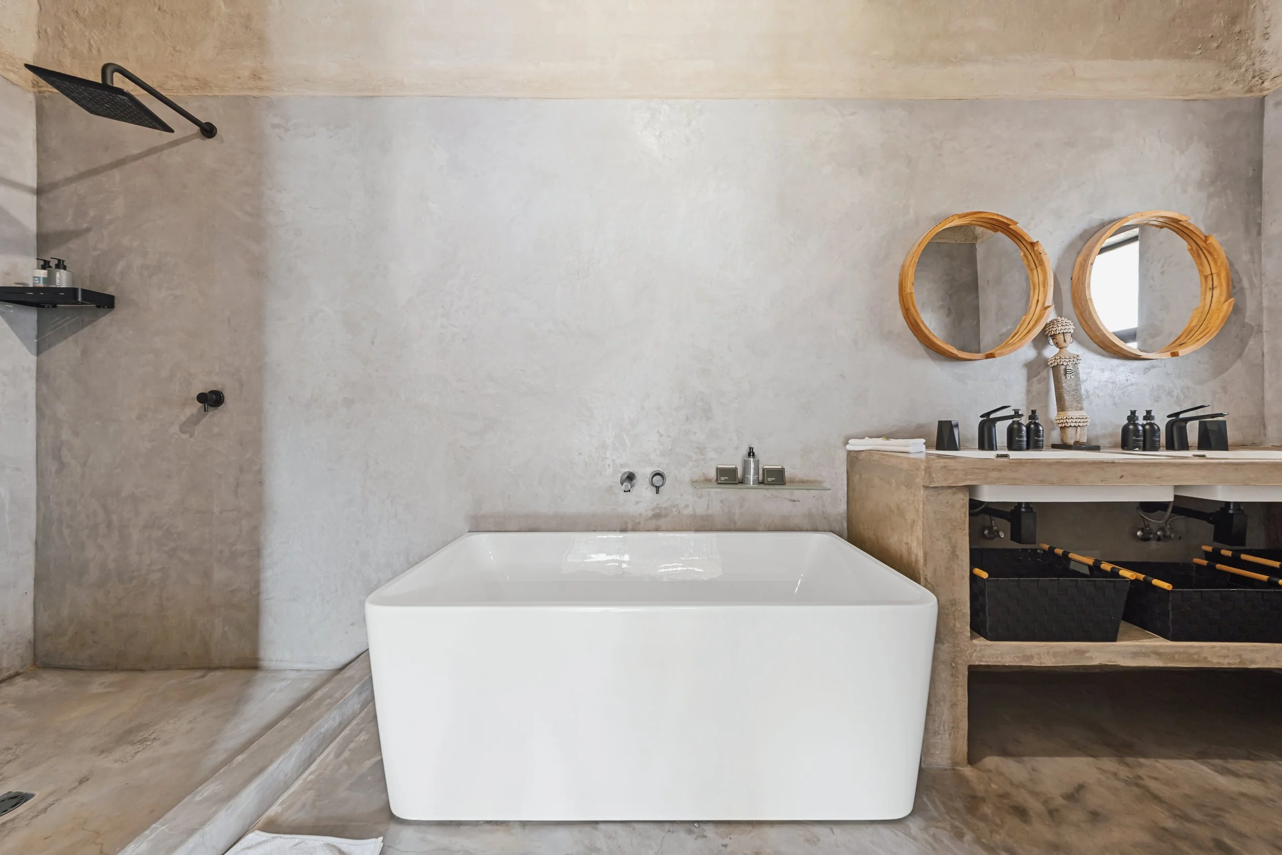 Modern bathroom with concrete walls and floor, featuring a large white bathtub, two circular wooden mirrors above a concrete vanity with black faucets and containers, a shelving unit with toiletries, and a black showerhead.