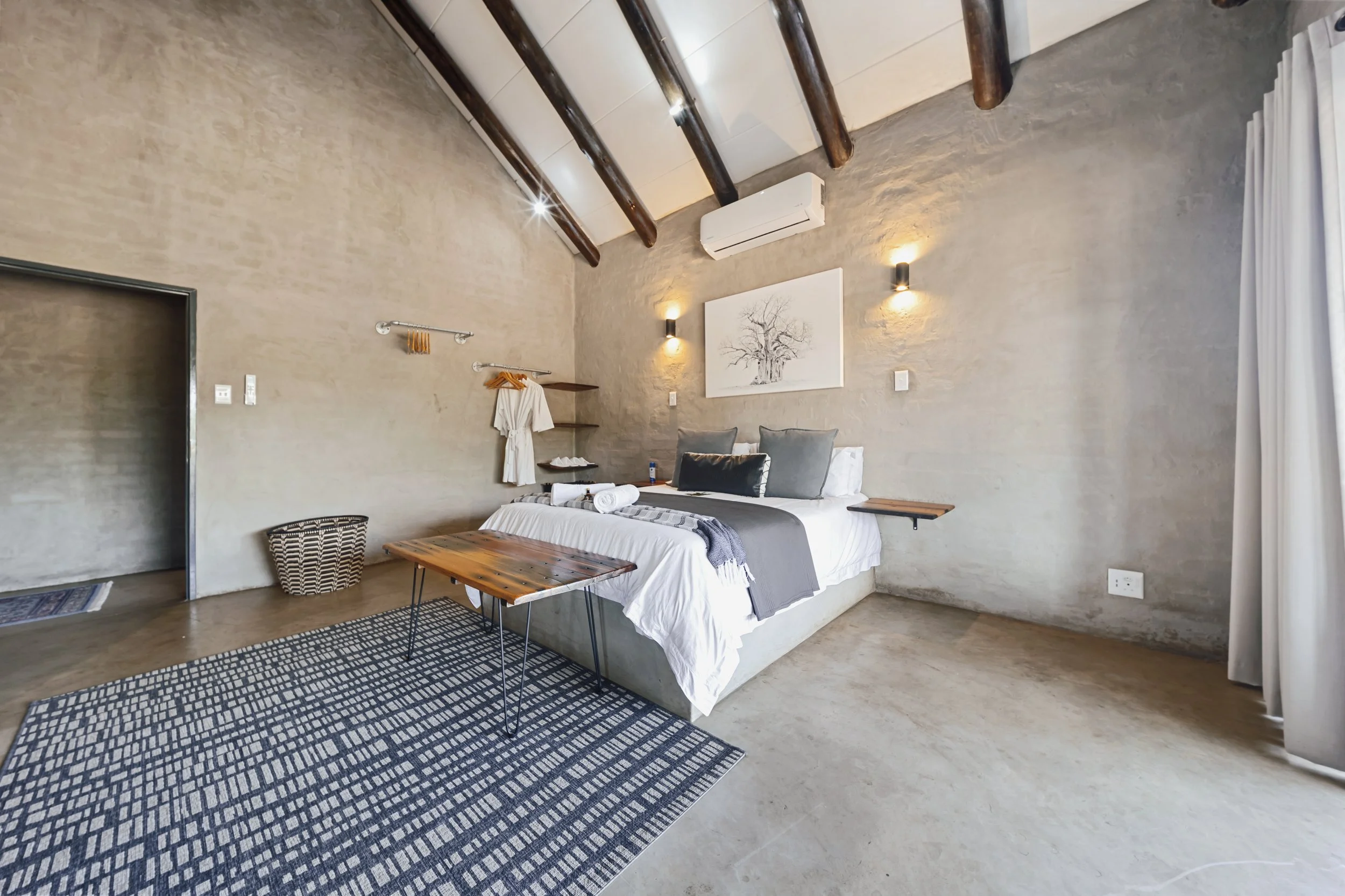 A bedroom with a bed, a wooden coffee table, and a patterned rug on a concrete floor. The walls are textured with beige paint, decorated with artwork and wall lights. There is an air conditioning unit on the wall, and a curtain covering a window.