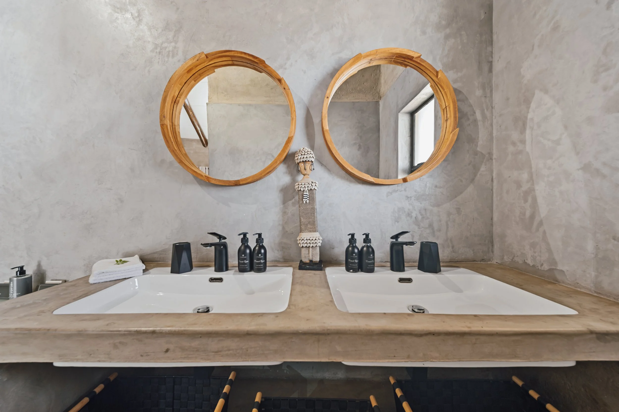 Double vanity bathroom sink with a beige countertop, two white sinks, and black fixtures. A decorative figure with shells is positioned between the sinks. Above the sinks are two round wooden-framed mirrors. On the beige countertop are bottles of soa