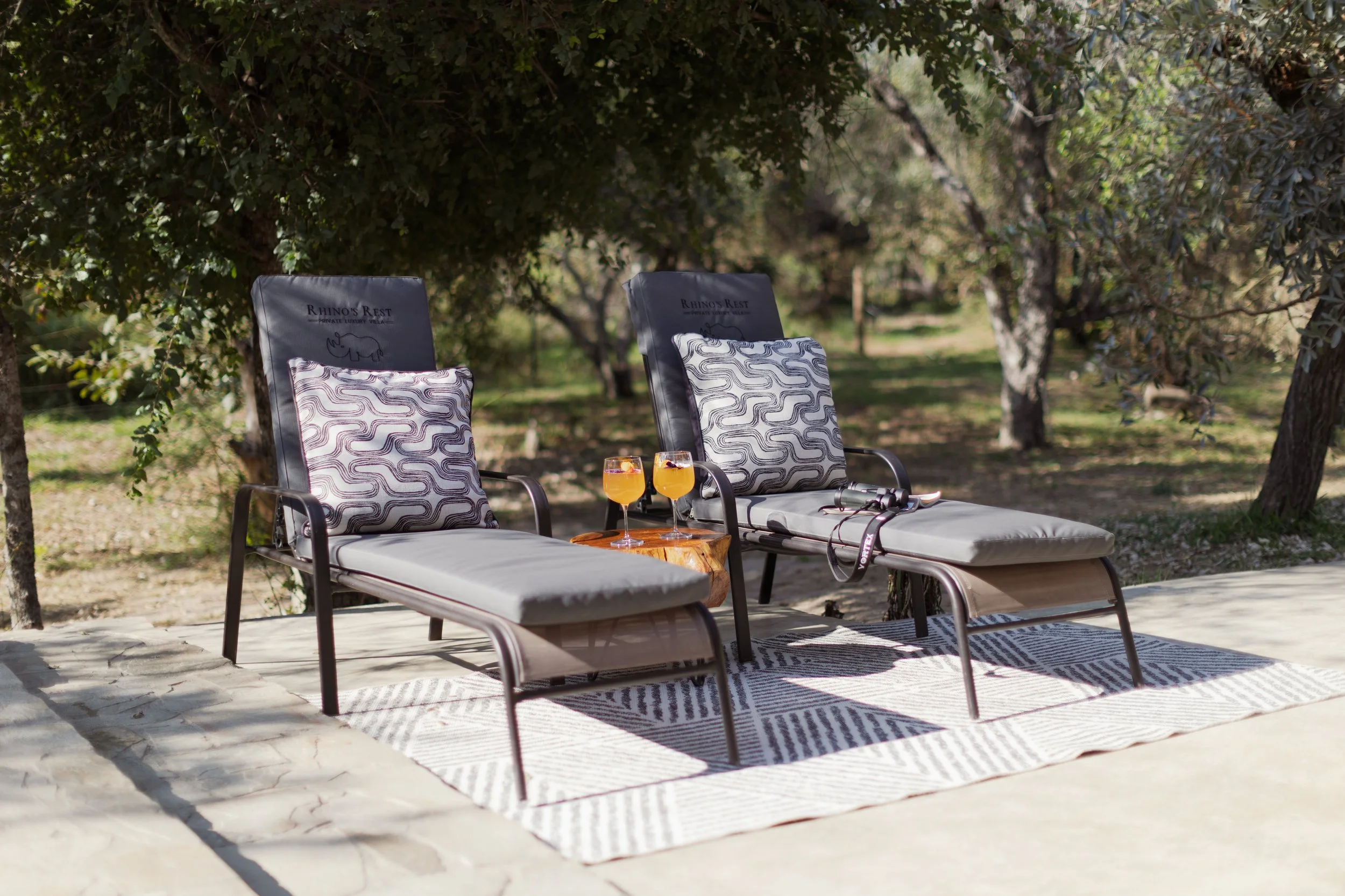 Two lounge chairs with patterned pillows set on a patio, two glasses of orange drinks with fruit garnish on a wooden table, surrounded by trees and greenery.