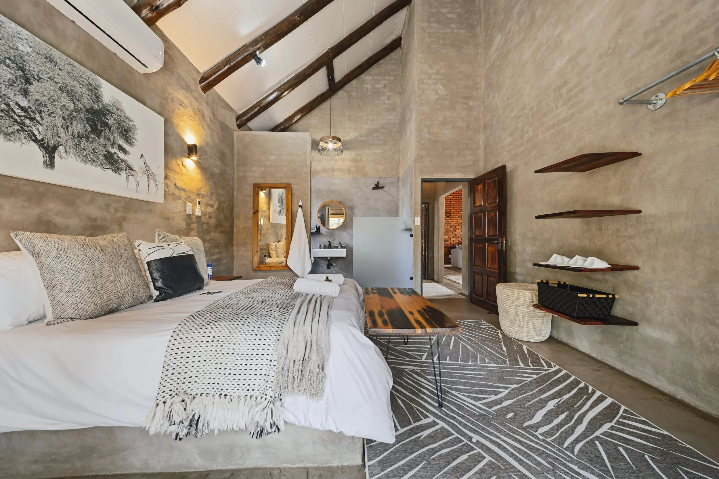 A cozy bedroom with a rustic aesthetic featuring a bed with pillows and a bed throw, a wooden coffee table, wall-mounted shelves, a large black and white wall art of a tree and giraffes, a mirror, towels, and a small sink area with a mirror. The ceil