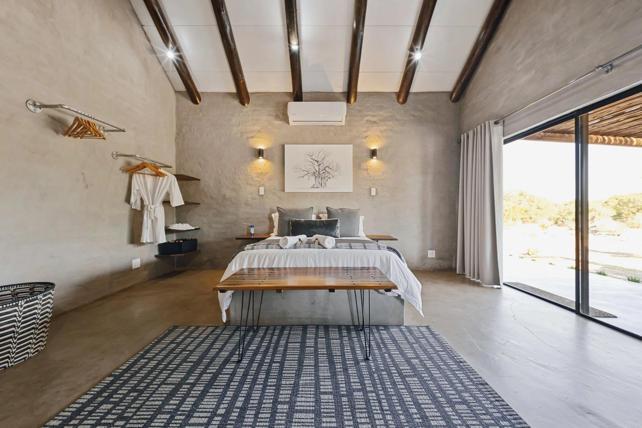 A bedroom with a bed, two wall-mounted lights, a framed tree picture, a window with sliding glass door and curtains, wood ceiling beams, and a woven rug.