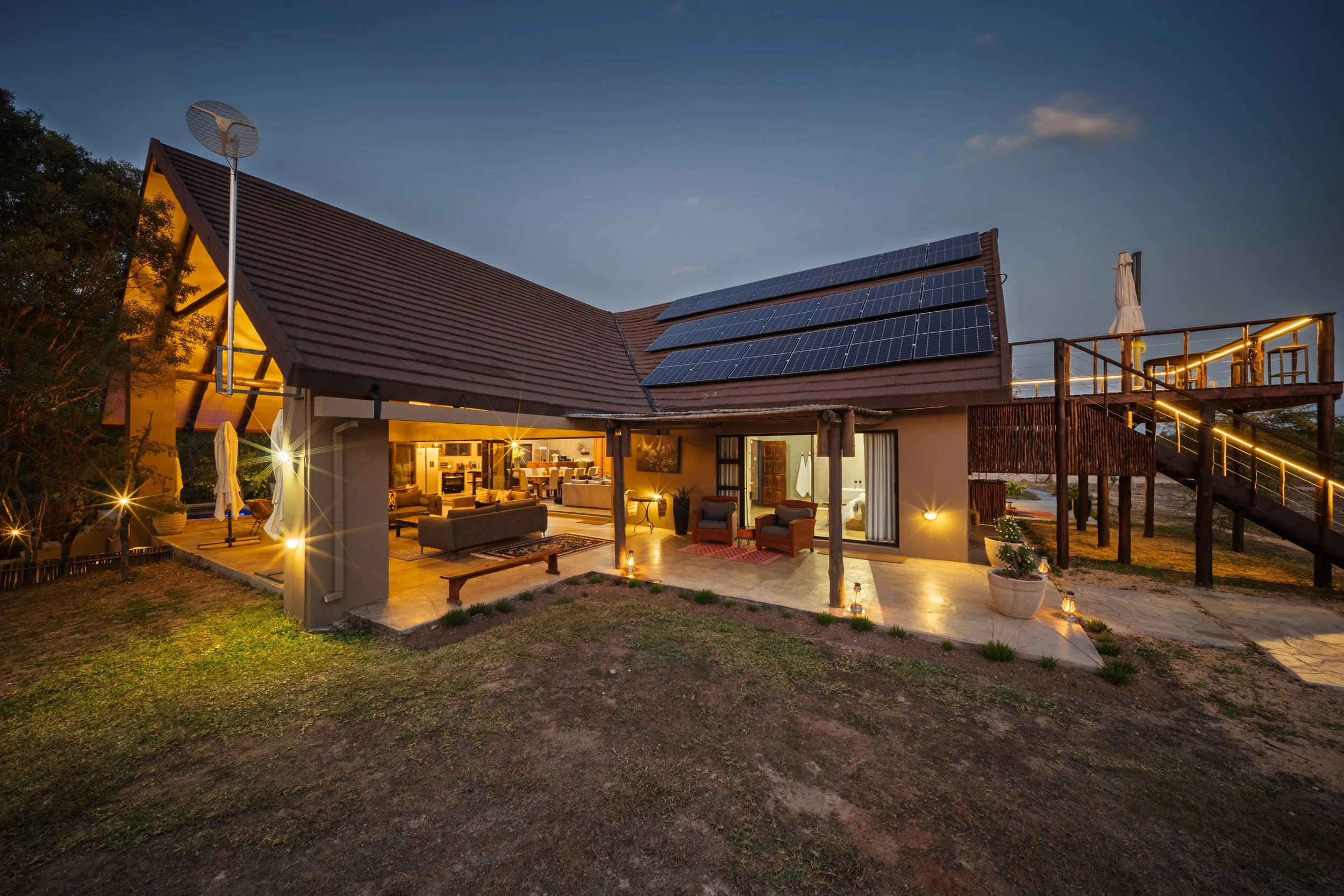A house with solar panels on the roof, an outdoor patio with furniture and string lights, a staircase with a white umbrella, and an evening sky with some clouds.