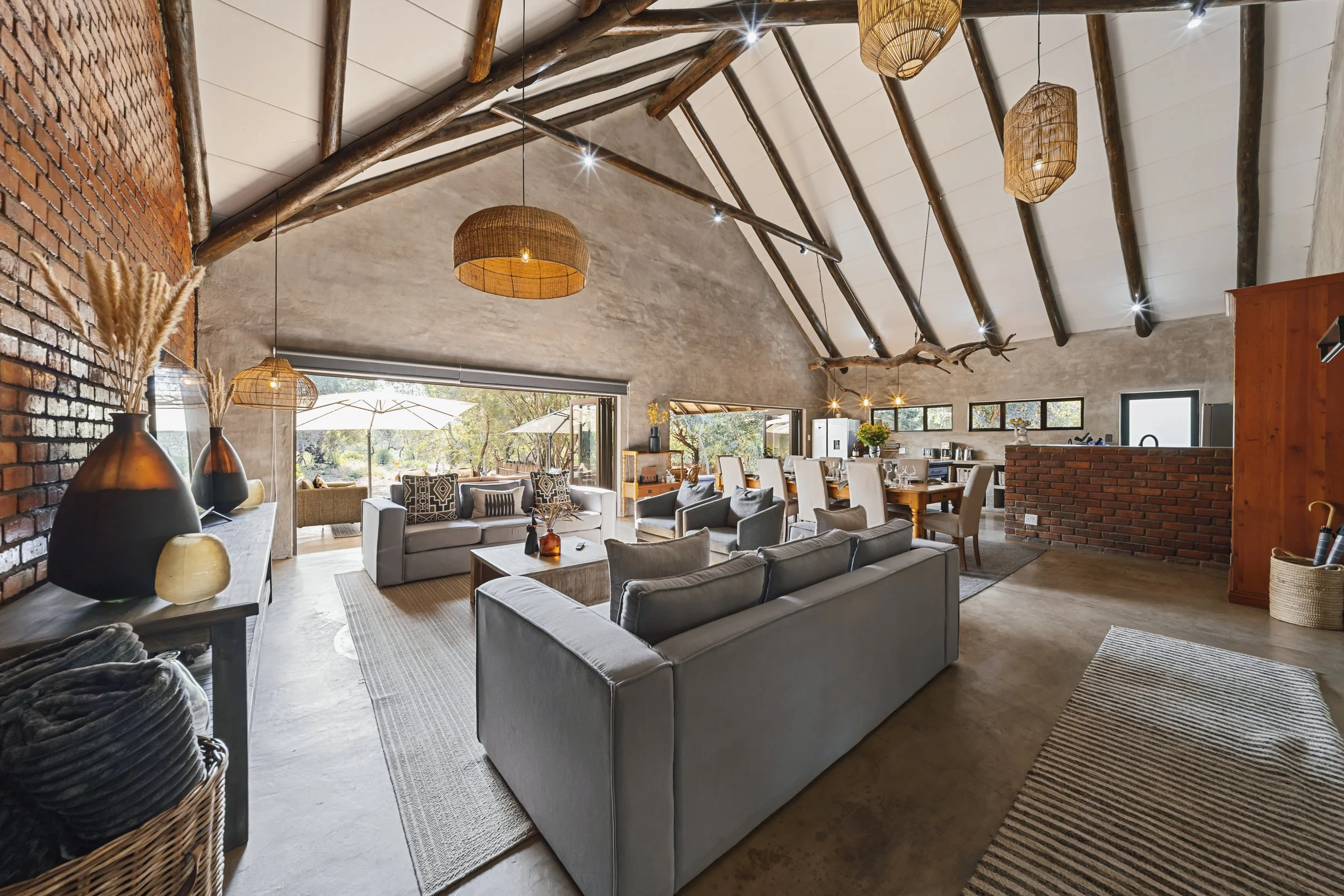 Open plan living and dining area with vaulted ceiling featuring exposed wooden beams and string lights. The room has modern and rustic decor, with large windows and glass doors providing natural light and outdoor views. There's a seating area with so