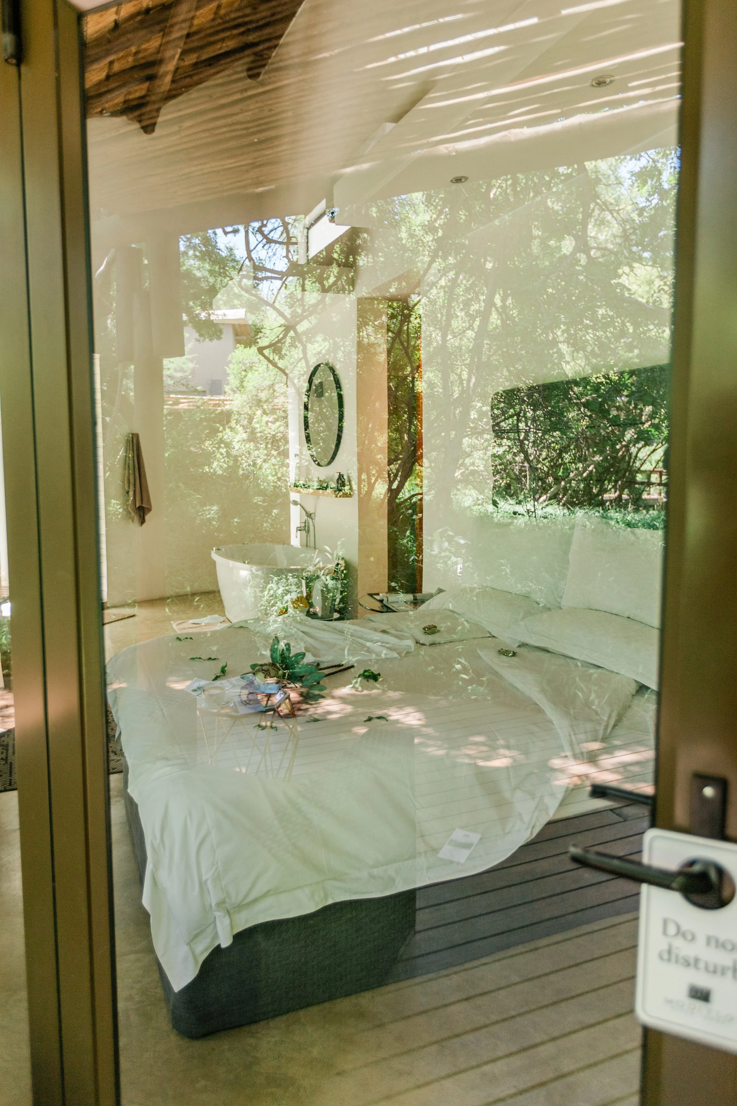 Reflected view of a bedroom with a bed, plants, and outdoor trees visible through a glass door