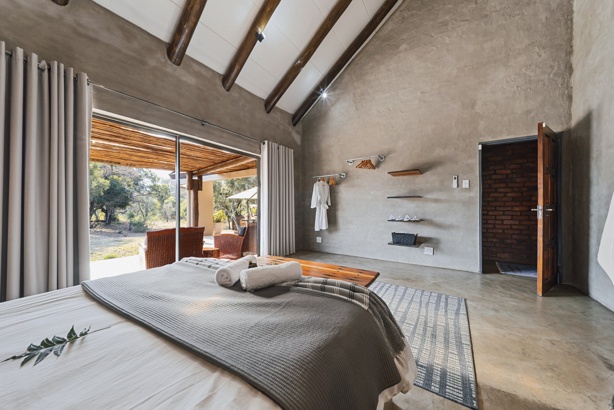 A bedroom with a bed, towels, and a view of a patio with outdoor chairs and trees outside.