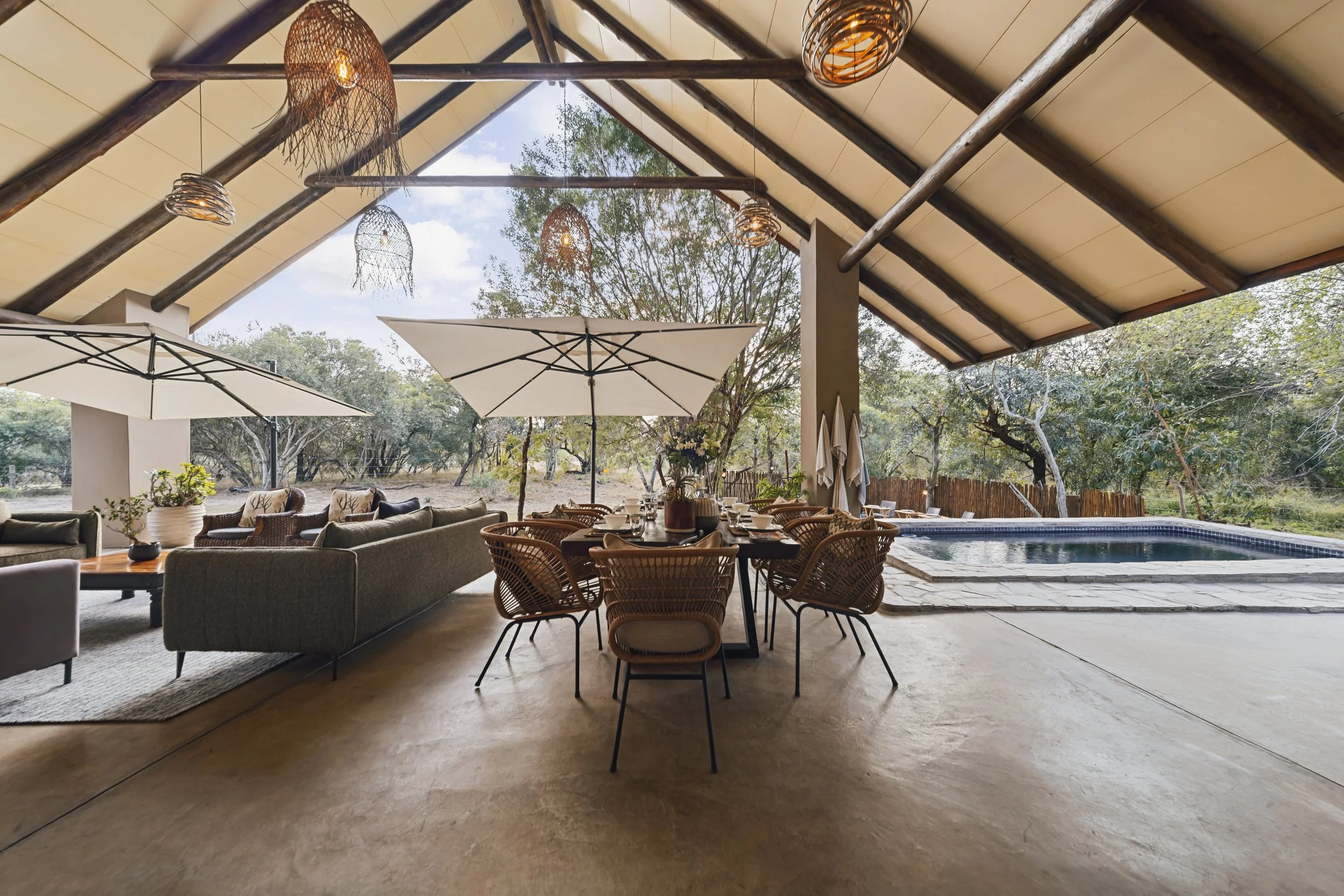 Indoor-outdoor living space with a ceiling featuring wooden beams and hanging lights, large windows with a view of trees, a pool, and a dining area with tables, chairs, and umbrellas.