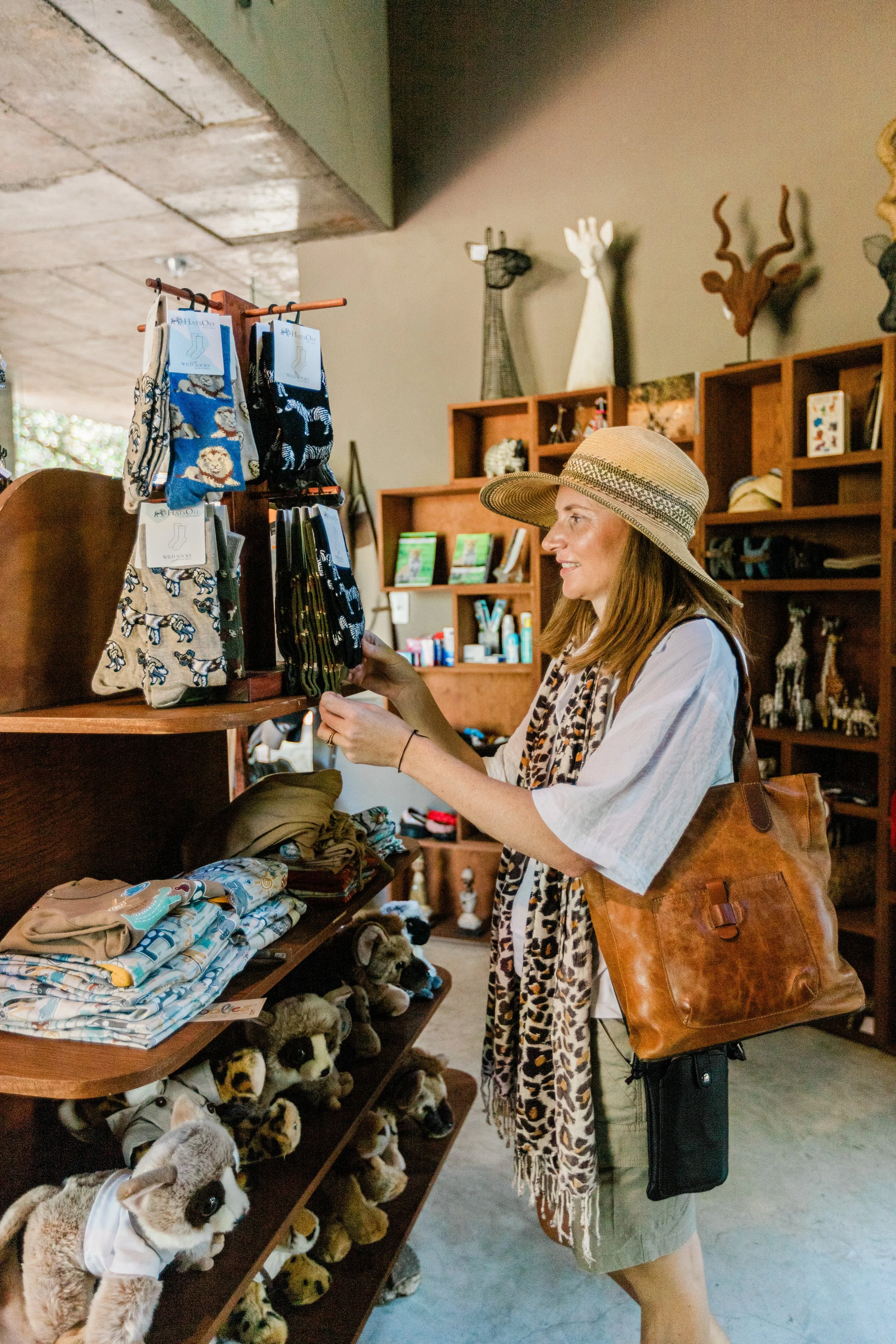 A woman wearing a leopard-print scarf shopping at a curio shop for gifts from South Africa. The store has wooden shelves with various items, including books and decorative objects, local crafts, safari clothing, toys and stuffed animals.
