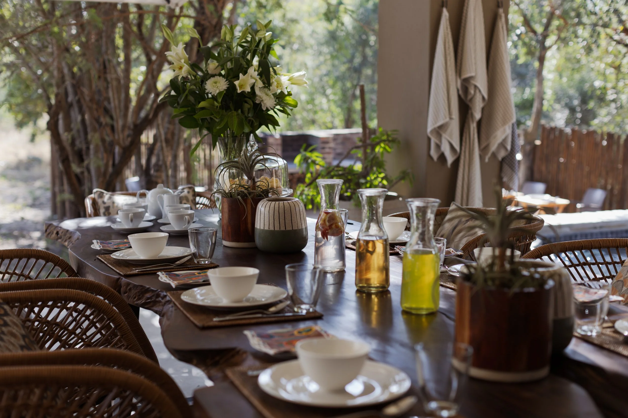 An outdoor dining table set with white cups, glass pitchers, and decorative vases, surrounded by wicker chairs with patterned cushions, under a tree with hanging towels or blankets on the side wall.