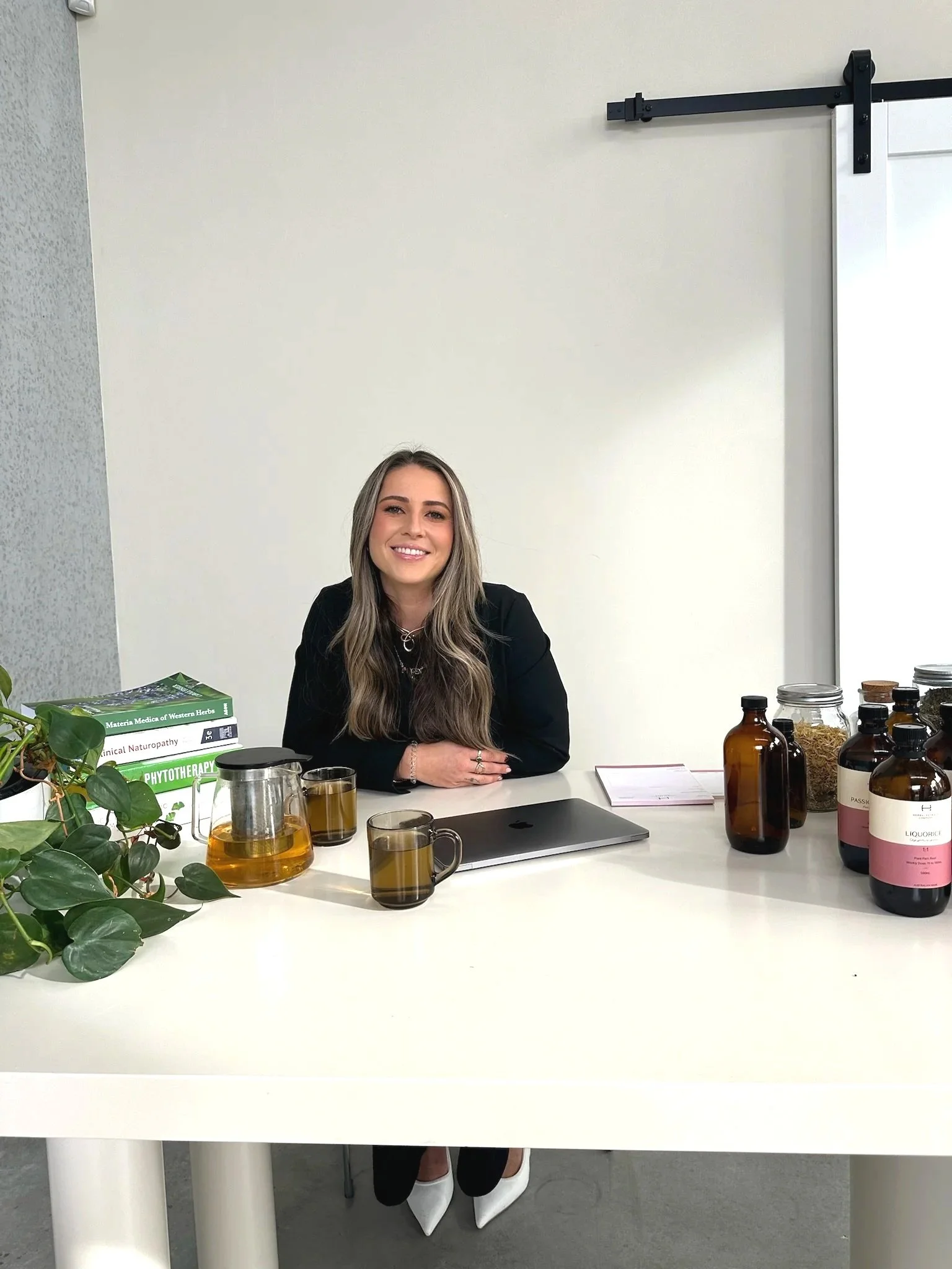 A woman sitting at a white desk with various bottles, jars, and books on herbal medicine, smiling at the camera.