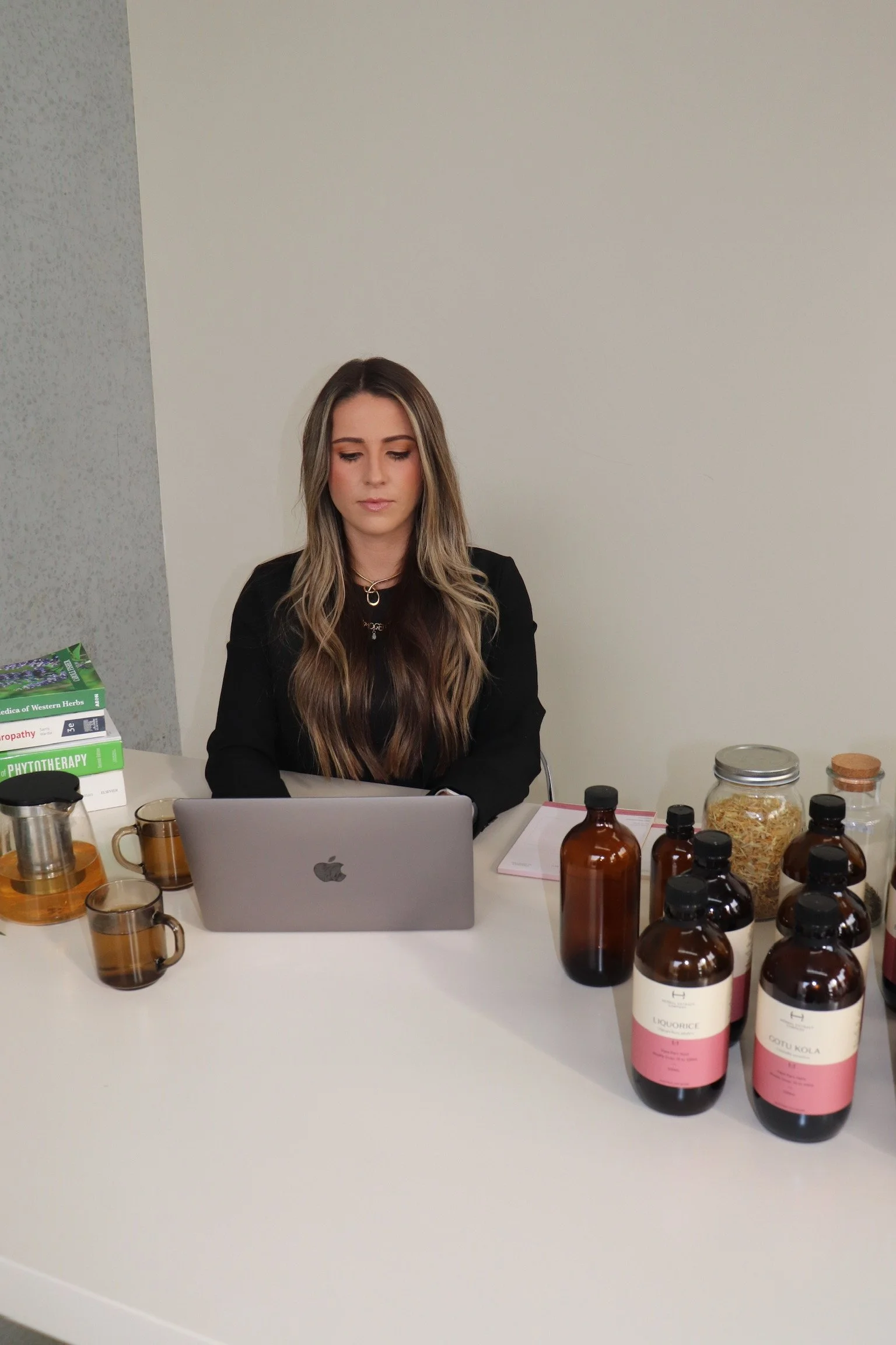 A woman with long wavy hair sitting at a white table, working on a laptop. The table has various bottles, jars, and teacups, with a stack of books in the background.