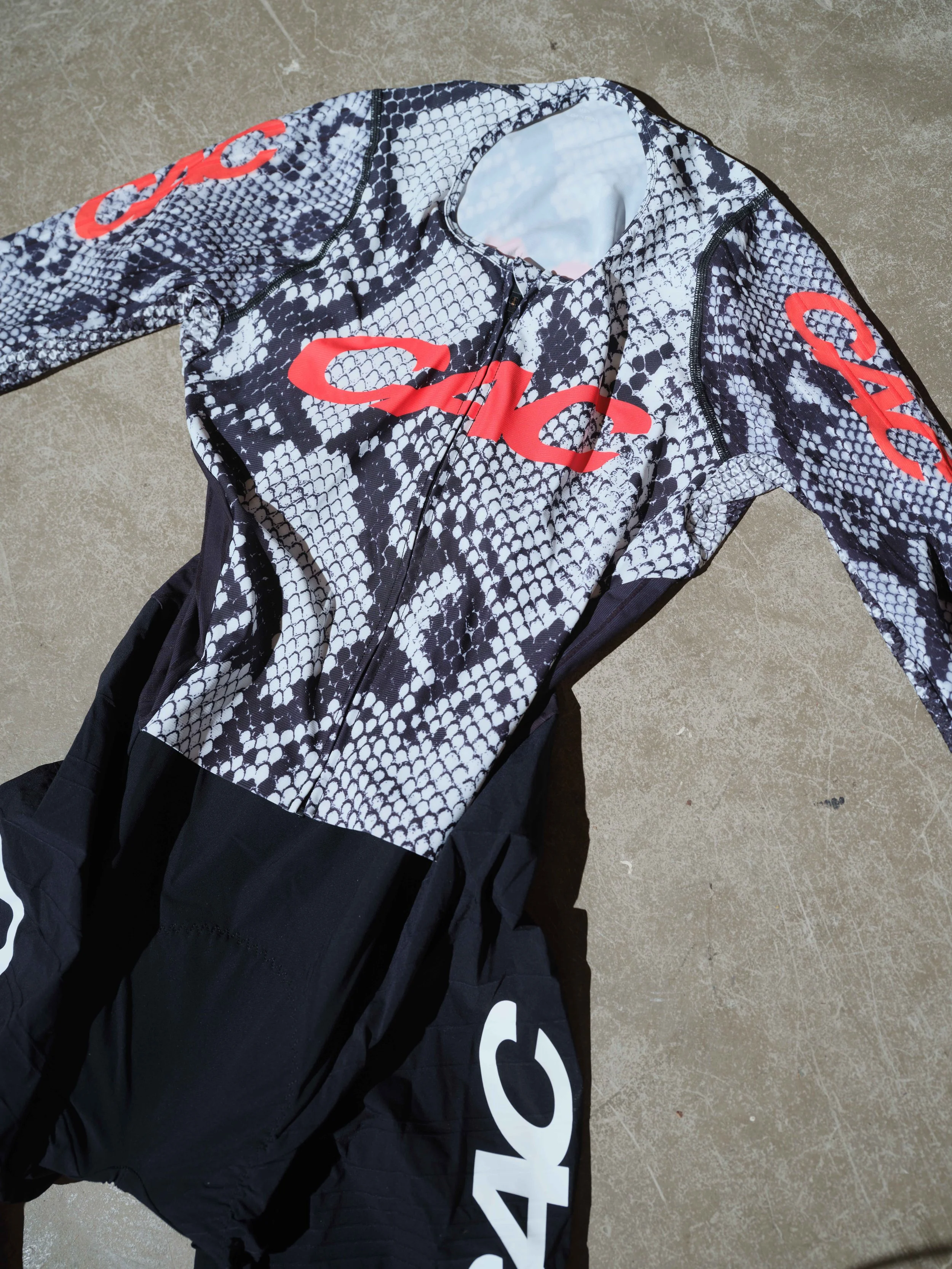 A long-sleeved cycling skin suit with a snakeskin pattern and red lettering, lying on a concrete surface.