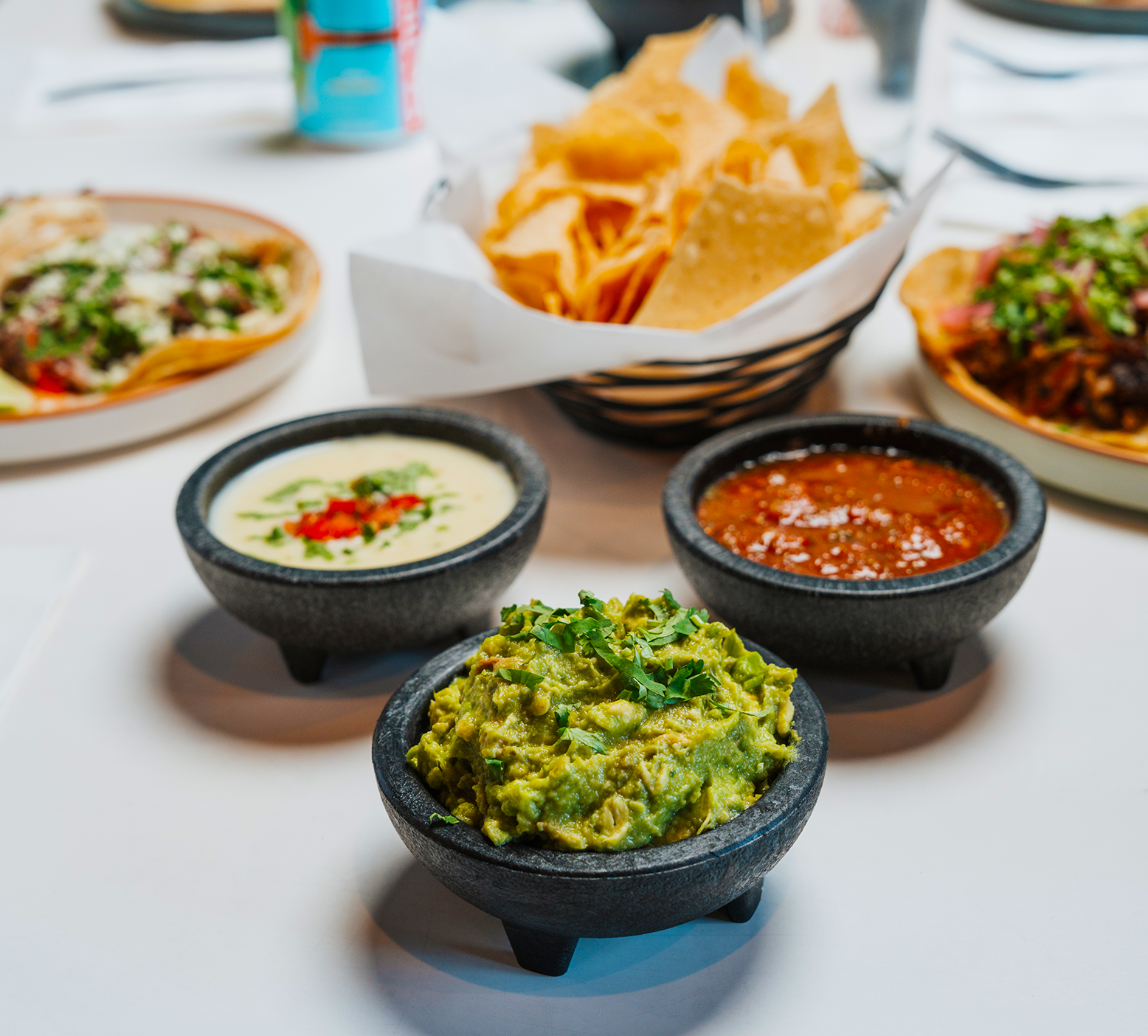 Table with bowls of guacamole, salsa, and sour cream, along with plates of tacos, tortilla chips, and Mexican dishes in the background.