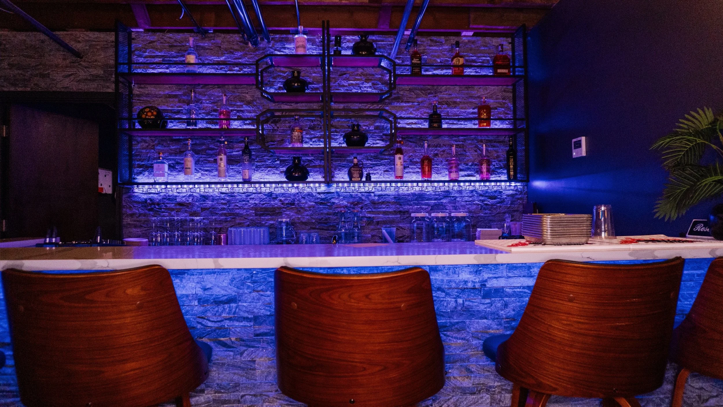 An illuminated bar area with a stone wall background, showcasing various bottles of alcohol on shelves with purple and blue lighting. There is a white marble counter with empty glasses and stacked trays, and four wooden barstools in front.