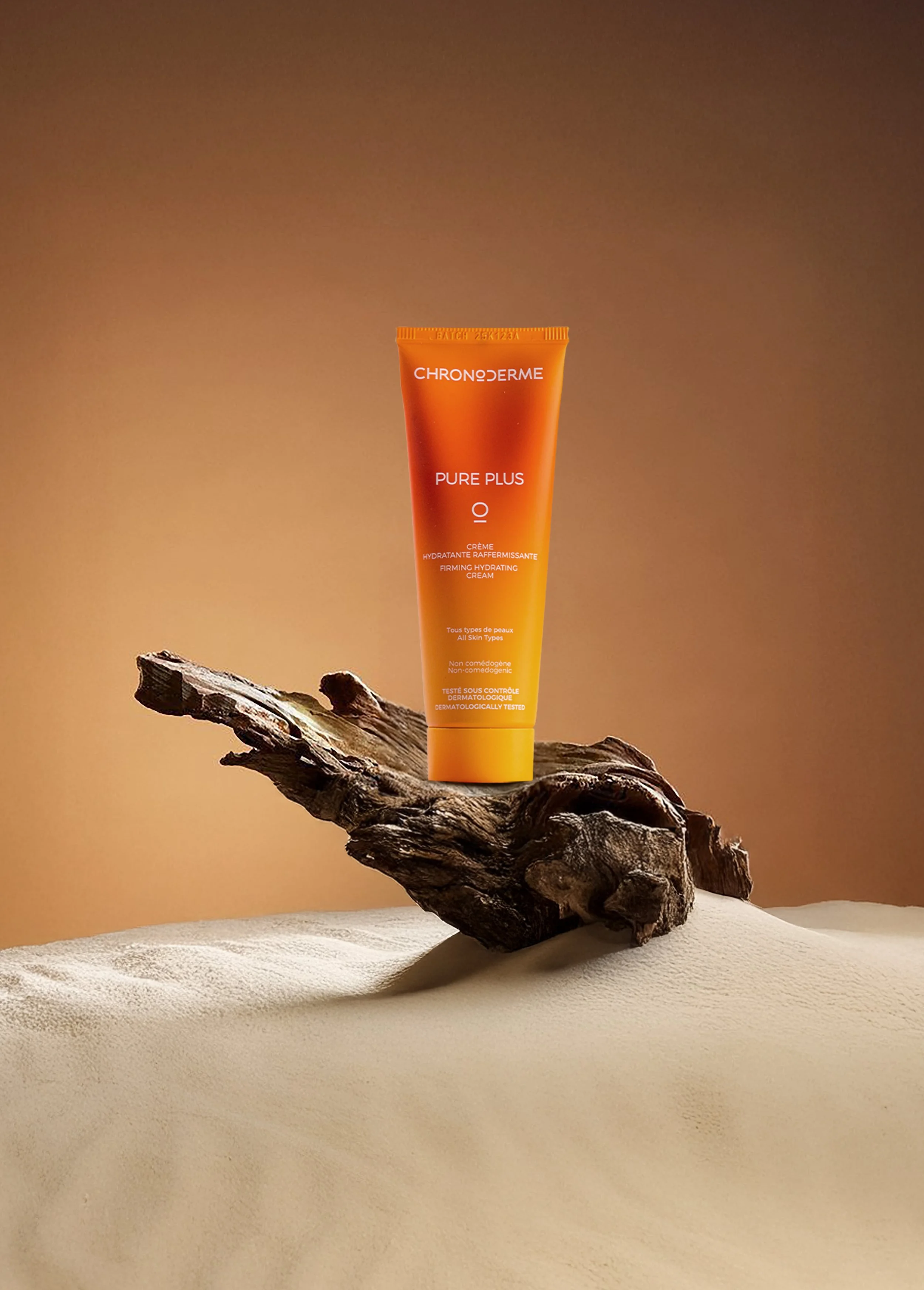 A tube of Chronoderme Pure Plus firming hydrating cream is placed on a piece of weathered wood, with a sandy surface in the foreground and a gradient brown background.