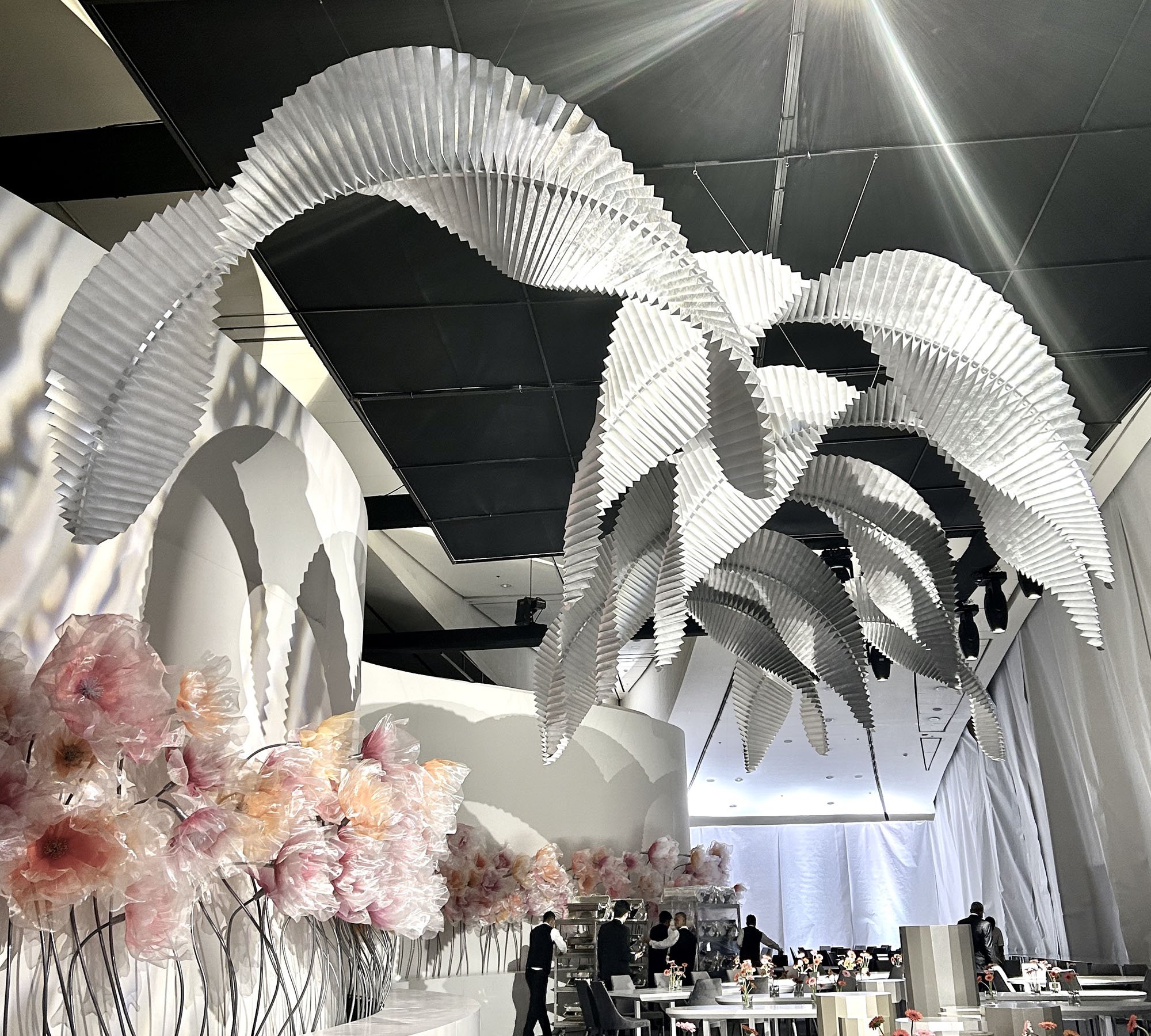 Large black ceiling with large white origami-style hanging sculpture resembling leaves, pink flowers in vases on tables, and people in a modern event space with white walls and curtains.