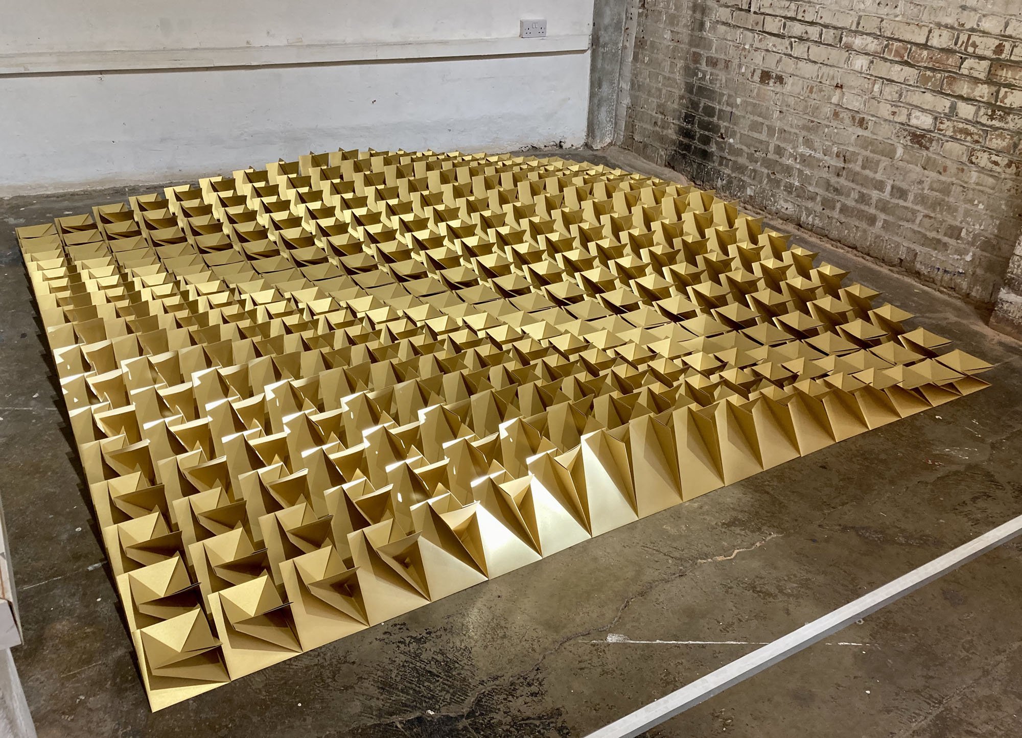 An art installation consisting of a large grid of gold-colored and gray paper or cardboard sculptures with folded, three-dimensional shapes, arranged on the floor in an industrial space with brick and concrete walls.
