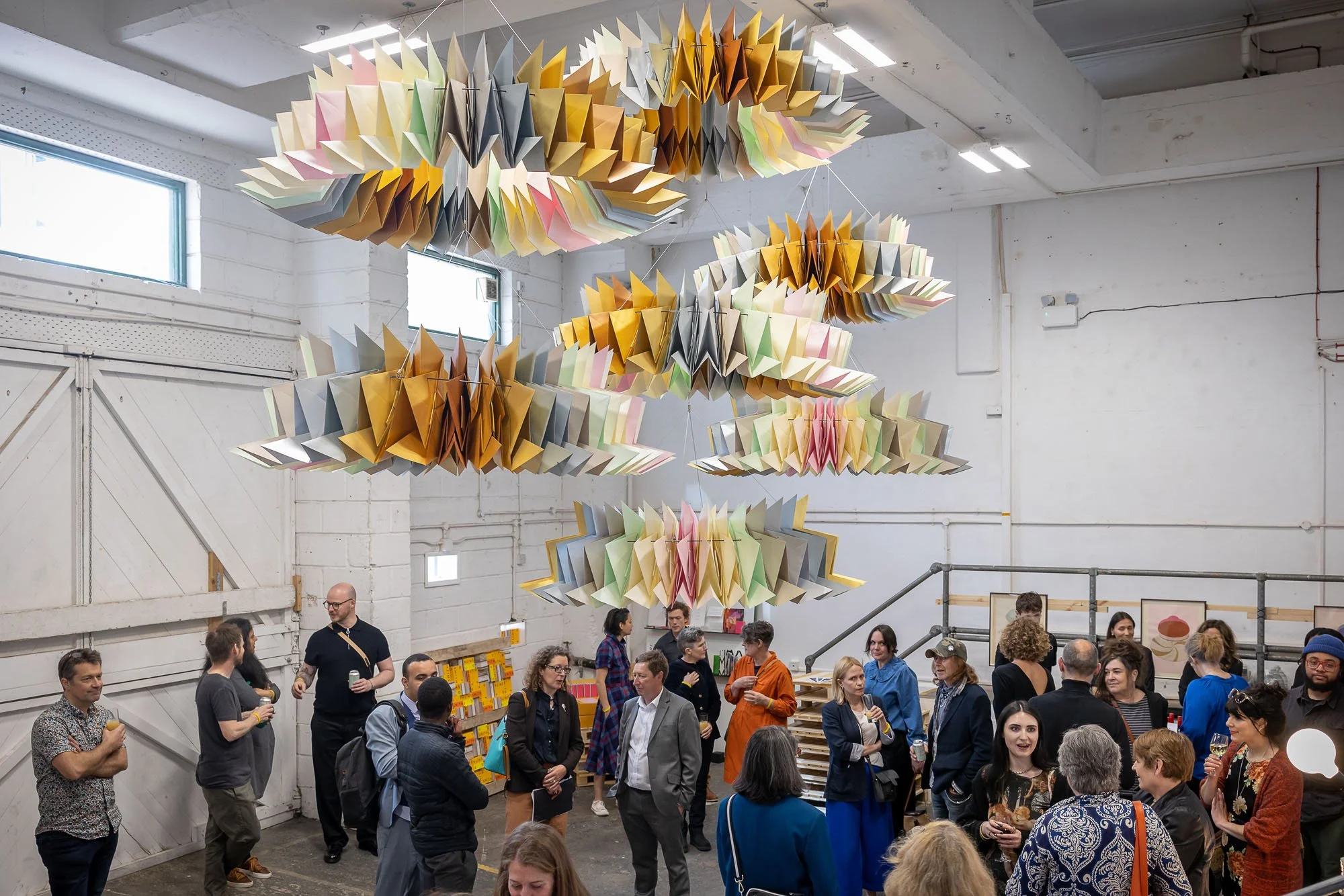 People attending an art exhibition in a warehouse-like space with white walls and large windows. The focal point is a large, colorful, abstract paper sculpture hanging from the ceiling, composed of layered, folded paper in pastel and neutral colors.