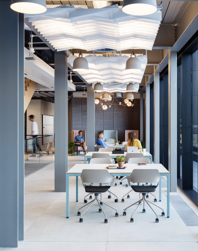 Modern office space with white desks, chairs, and potted plants, illuminated by large, white, textured ceiling lights and pendant lights. People are working and conversing in the background, with glass windows on the right.