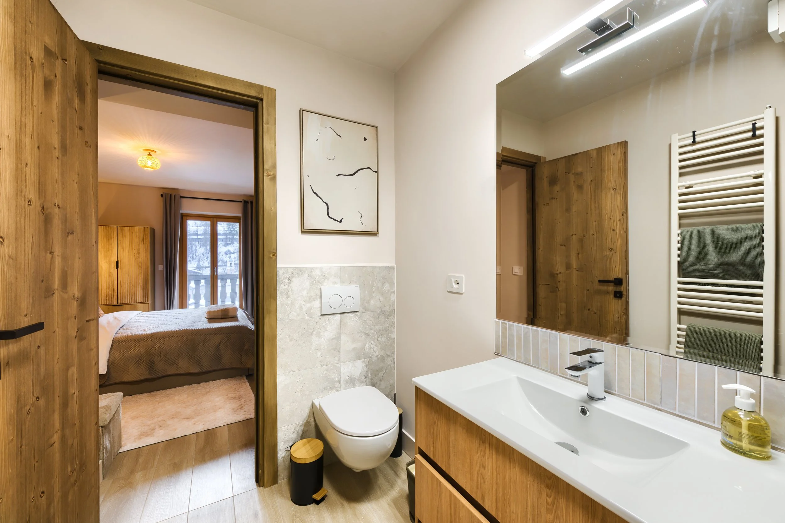 Modern bathroom with white sink and wooden cabinetry, mirror, towel warmer, and a view into a bedroom with a bed, wooden furniture, and balcony door, a luxurious retreat, an exclusive retreat, a refined mountain retreat, a premium alpine retreat.
