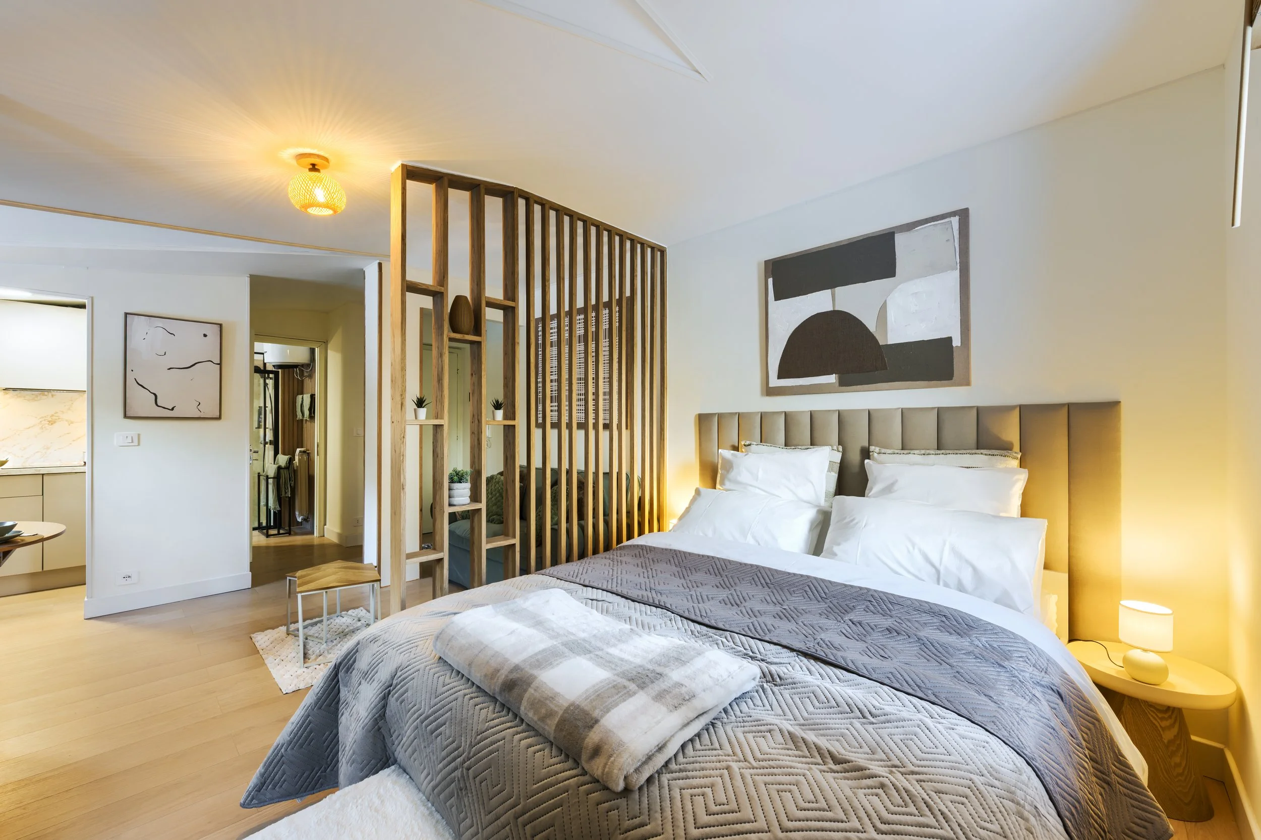 A modern bedroom with a bed that has a beige headboard, white pillows, and a gray quilt. A small lamp sits on a wooden nightstand, and abstract black and white artwork hangs above the bed. There is a wooden partition with small potted plants separati