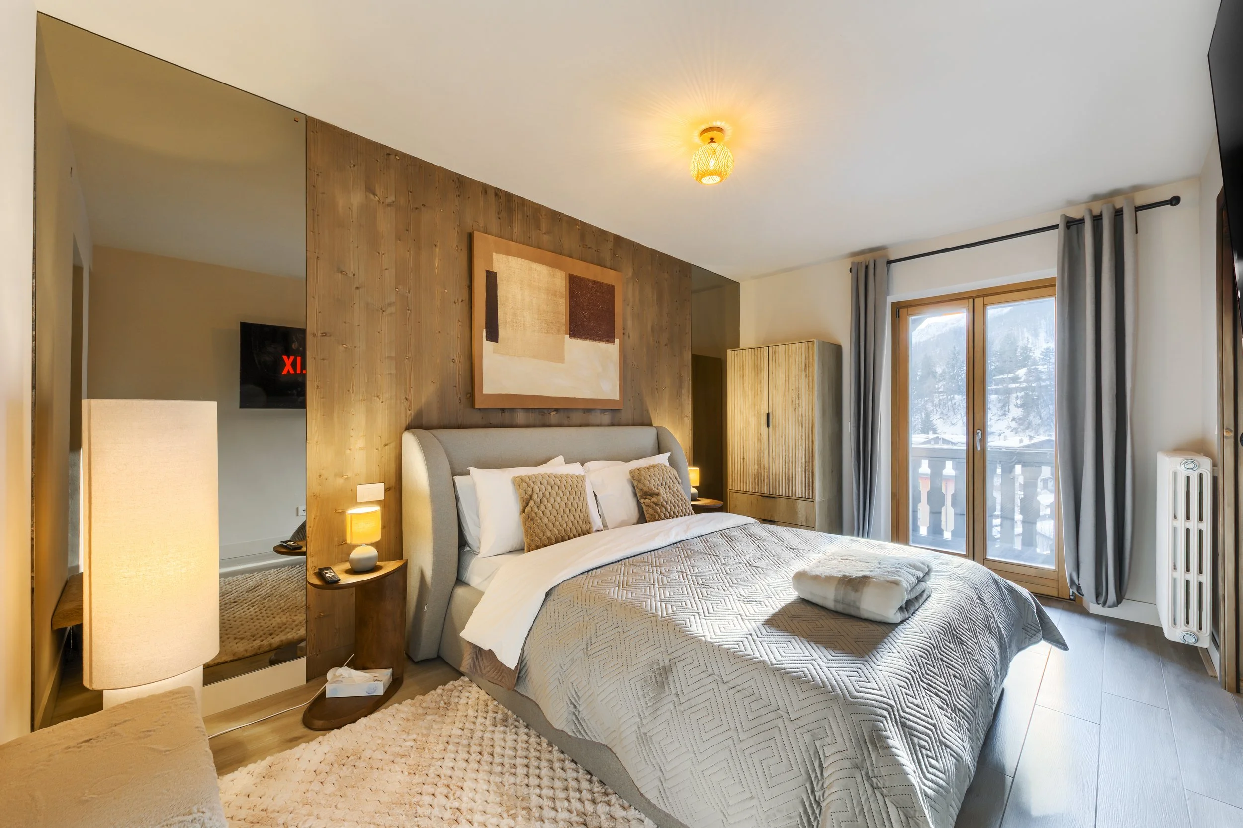Cozy bedroom with a large bed, beige headboard, wooden accents, abstract artwork, sliding glass door with curtains, bedside lamps, and winter landscape outside. a luxurious retreat, an exclusive retreat, a refined mountain retreat, premium alpine.