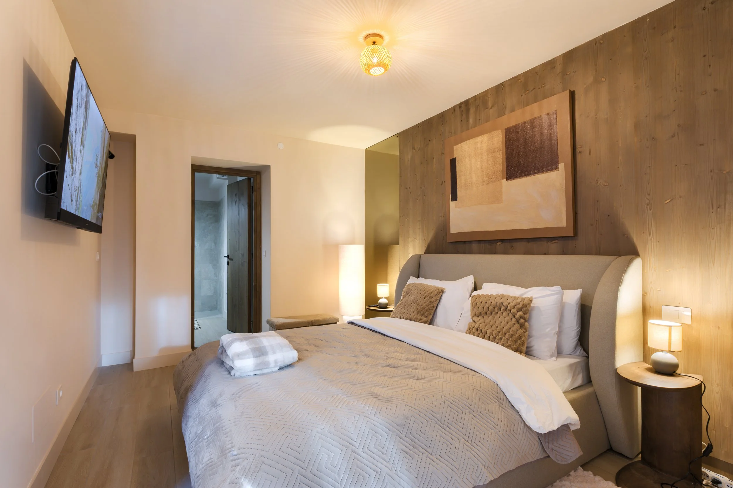 A cozy bedroom with a large bed, beige and brown pillows, a wooden headboard, and two bedside lamps. A flat-screen TV is mounted on the wall opposite the bed, and there is a doorway leading to a bathroom. a luxurious retreat, an exclusive retreat.